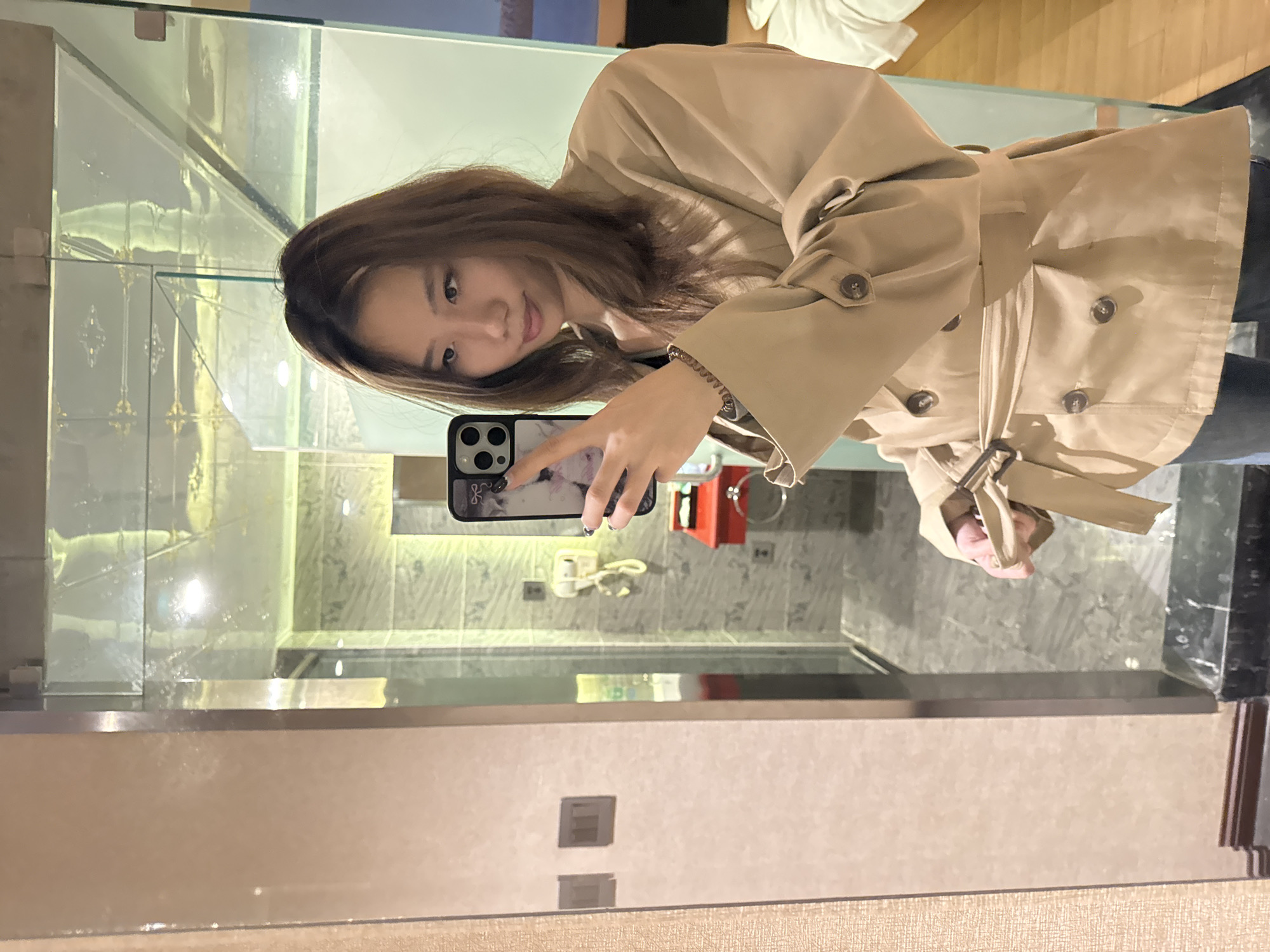 The image displays a mirror selfie of a young woman, likely East Asian, taken indoors within a modern setting, specified as Ganzhou, China. The woman is positioned with her body angled, looking towards her reflection and holding a smartphone in her right hand to capture the image.
She has long, straight brown hair and a subtle, pleasant expression. She is dressed in a light beige trench coat, characterized by a belted waist, visible buttons on the front, and adjustable cuffs, one of which shows a button closure. A dark, spiral-shaped hair tie or bracelet adorns her left wrist. The smartphone she holds features a prominent triple-lens camera array, indicative of an iPhone Pro model, and has a light-colored, patterned case. The faint Apple logo is visible on the phone's back.
The background, largely composed of her reflection, reveals a contemporary interior. Elements include a clear glass partition, possibly for a shower, and light-colored, veined wall tiles that resemble marble. Below the tiles, a sleek counter or vanity surface is visible. To the right of the woman's phone, a red, wall-mounted rectangular object, possibly a dispenser or storage unit, is reflected. Electrical outlets or switches can also be seen on the wall. The room is brightly lit, suggesting either strong ambient daylight or well-placed artificial lighting. No distinct, legible text beyond the Apple logo on the phone is apparent in the image.