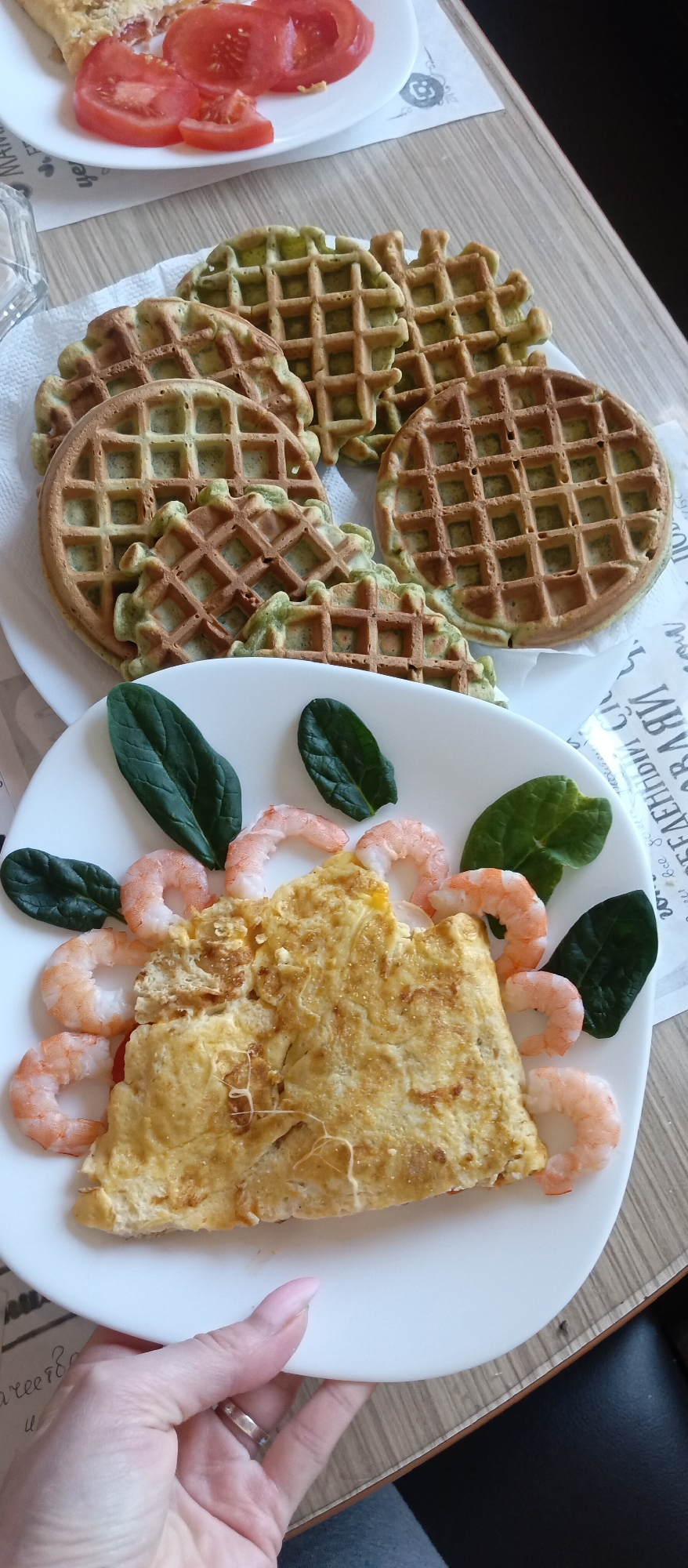 The image displays a delectable breakfast or brunch spread set on a light-colored wooden table. In the foreground, a hand, adorned with light pink nail polish and a silver ring, holds a white ceramic plate. This plate features a fluffy, golden-yellow omelet, elegantly surrounded by several cooked pink shrimp and vibrant, fresh dark green spinach leaves. Melted cheese strands are visible on the omelet, adding to its appetizing appearance.
In the mid-ground, resting on another white plate, is a collection of unique greenish waffles. These waffles exhibit a classic grid pattern, with some being perfectly round and others having more intricate, possibly heart-shaped, designs. Their distinct green tint suggests the incorporation of ingredients like spinach or matcha. Partially visible at the top left, on a small white plate, are several bright red slices of fresh tomato, possibly accompanying a pastry or savory dish.
Beneath the waffle plate, a white paper or napkin with black text is visible. The text appears to be in Cyrillic, with discernible words and fragments such as "ЗДРАВИ" (meaning "healthy" or "hello" in plural), "СЛАВИ" (possibly part of "congratulations"), "мам" (mom), and "Все" (all/everything). This suggests a decorative or newsprint paper, possibly conveying a message of health or well-wishes.
The scene depicts a carefully prepared meal, likely enjoyed indoors during the daytime. The combination of savory omelet with shrimp and spinach, alongside the intriguing green waffles and fresh tomatoes, presents a balanced and appealing dining experience.
