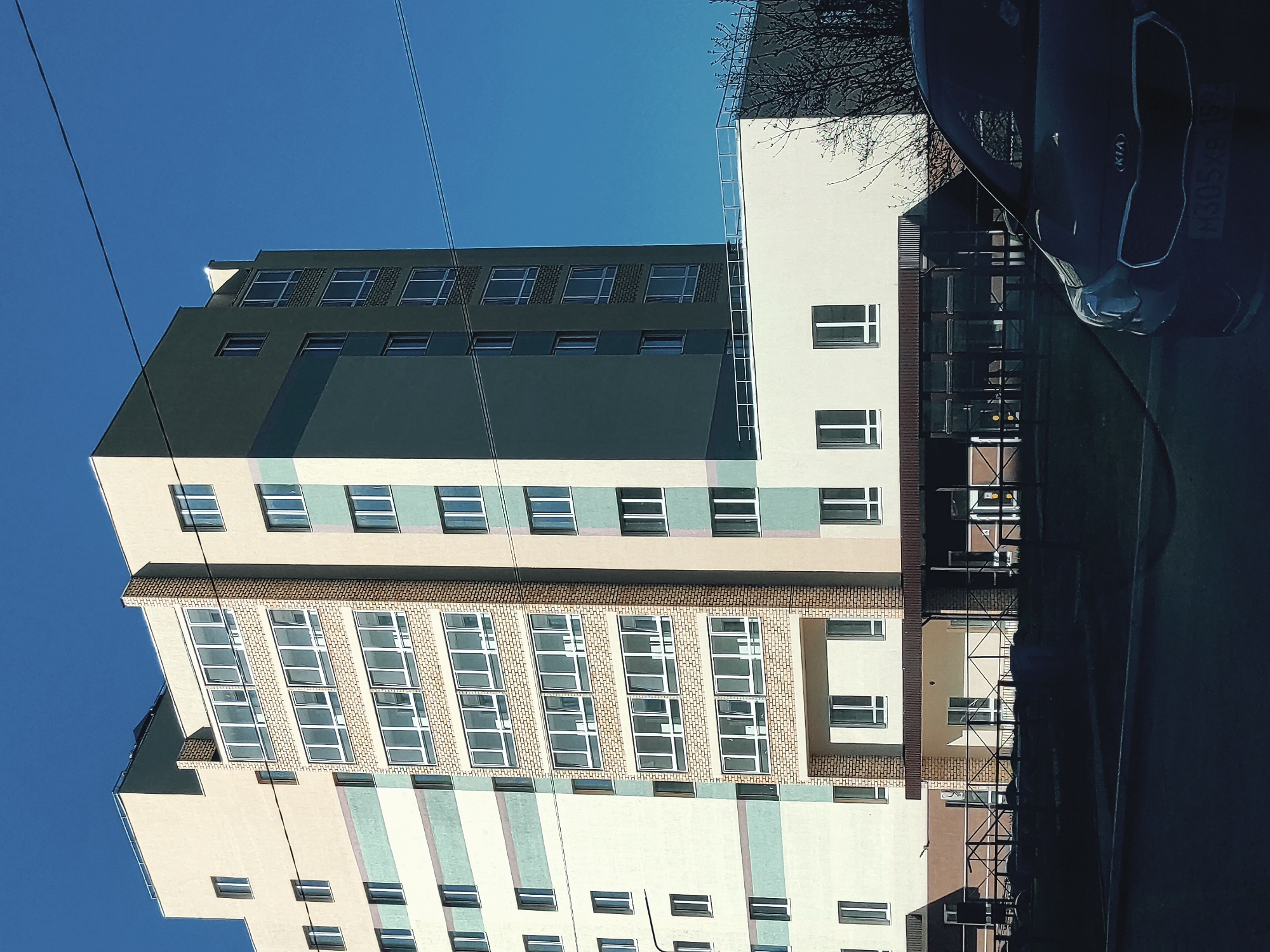 Here's a description of the image:
**Scene:** A medium shot photograph of a multi-story residential building under a clear blue sky. The building features a varied color scheme with segments of beige, light green, and a darker gray near the top. The building is punctuated with rows of windows, some framed by brick. An electricity cable runs across the view.
**Objects:** A dark-colored Kia car is partially visible in the right of the frame. A sidewalk and a section of black metal railing are also visible.
**Location:** Based on the license plate on the car (M305AC159), the location is likely Chaikovsky, Russia.
