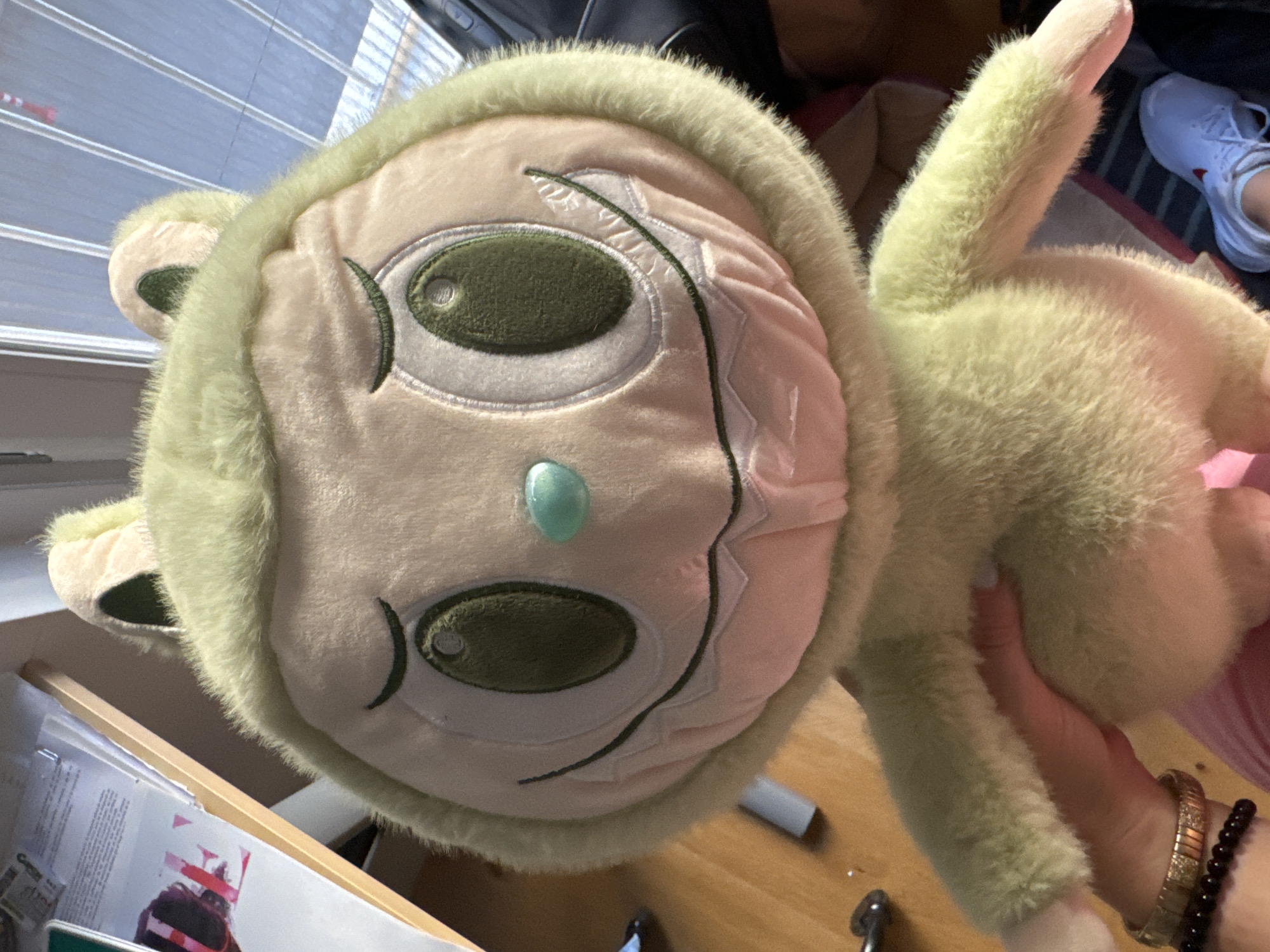 A light green plush toy with large, dark green eyes and a light pink face is held by a person's hand. The toy has a furry texture and a hood with light pink ears. Its mouth is depicted as a wide smile with jagged, white teeth and a dark line representing the lips. A small, light blue nose is visible. The background shows a wooden surface, papers, and a window with blinds. The person holding the toy is wearing a pink garment and a bracelet on their wrist. In the lower right portion of the image, a white sneaker is visible, suggesting the presence of another person. The overall scene appears to be indoors, possibly in a home or office environment. There is no indication of the city of Murcia, Spain in the provided image.