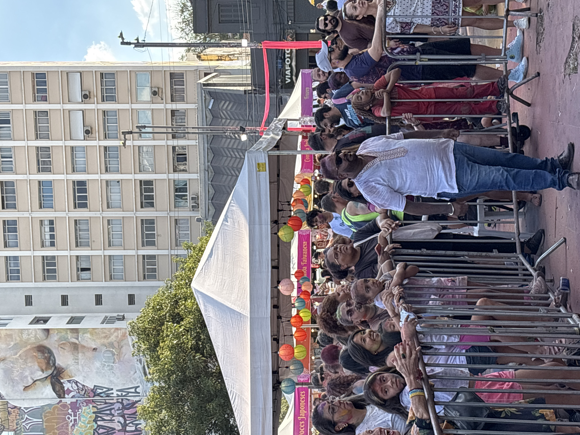 Captured from a low angle, the image shows a densely packed crowd of people behind a metal barricade, likely at a public event. A large white tent structure is visible on the left, adorned with strings of colorful lanterns. Banners with text that includes "Doces Japoneses" and "Comida Taiwanesa" hang from the tent. The background includes a multi-story building with numerous windows, and a mural or graffiti art is visible on a wall. Overhead, a red banner with "VIAFOTO" is visible. The crowd consists of people of varying ages and ethnicities, some leaning against the barricade, others looking toward the event. The setting appears to be a street in São Paulo, Brazil.