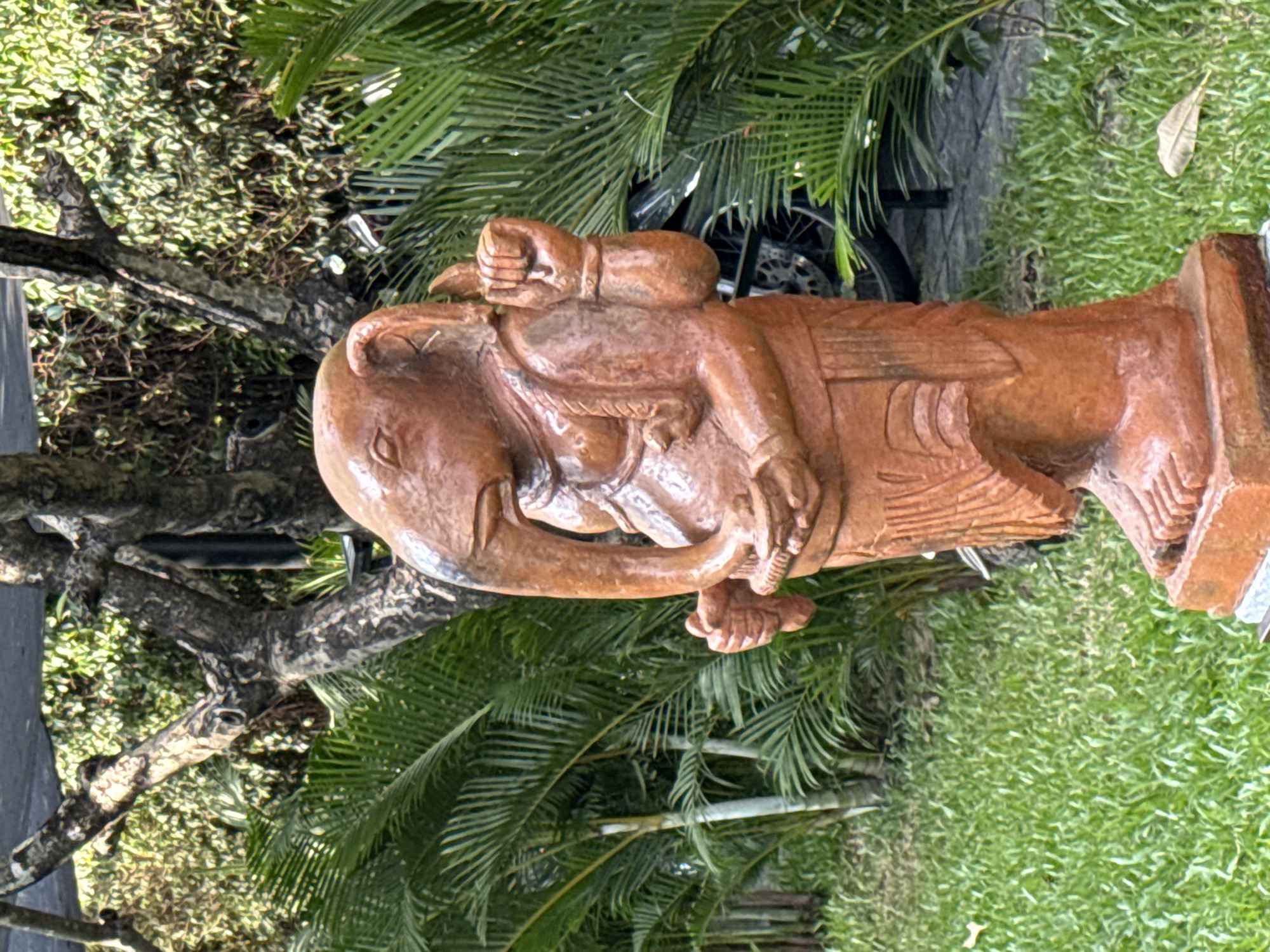 The image shows a statue of Ganesha, a Hindu deity, made of terracotta. The statue is positioned outdoors on a patch of green grass, with palm trees and other foliage in the background. The statue is a vibrant reddish-brown color and depicts Ganesha in a standing posture, holding various objects in his hands. The details of the statue, such as its multiple arms and elephant head, are clearly visible. The overall scene suggests a serene and tranquil environment, possibly a garden or a courtyard in a temple or a residential area. The background hints at a tropical climate, with lush greenery and sunlight filtering through the trees. There are no people visible in the image.