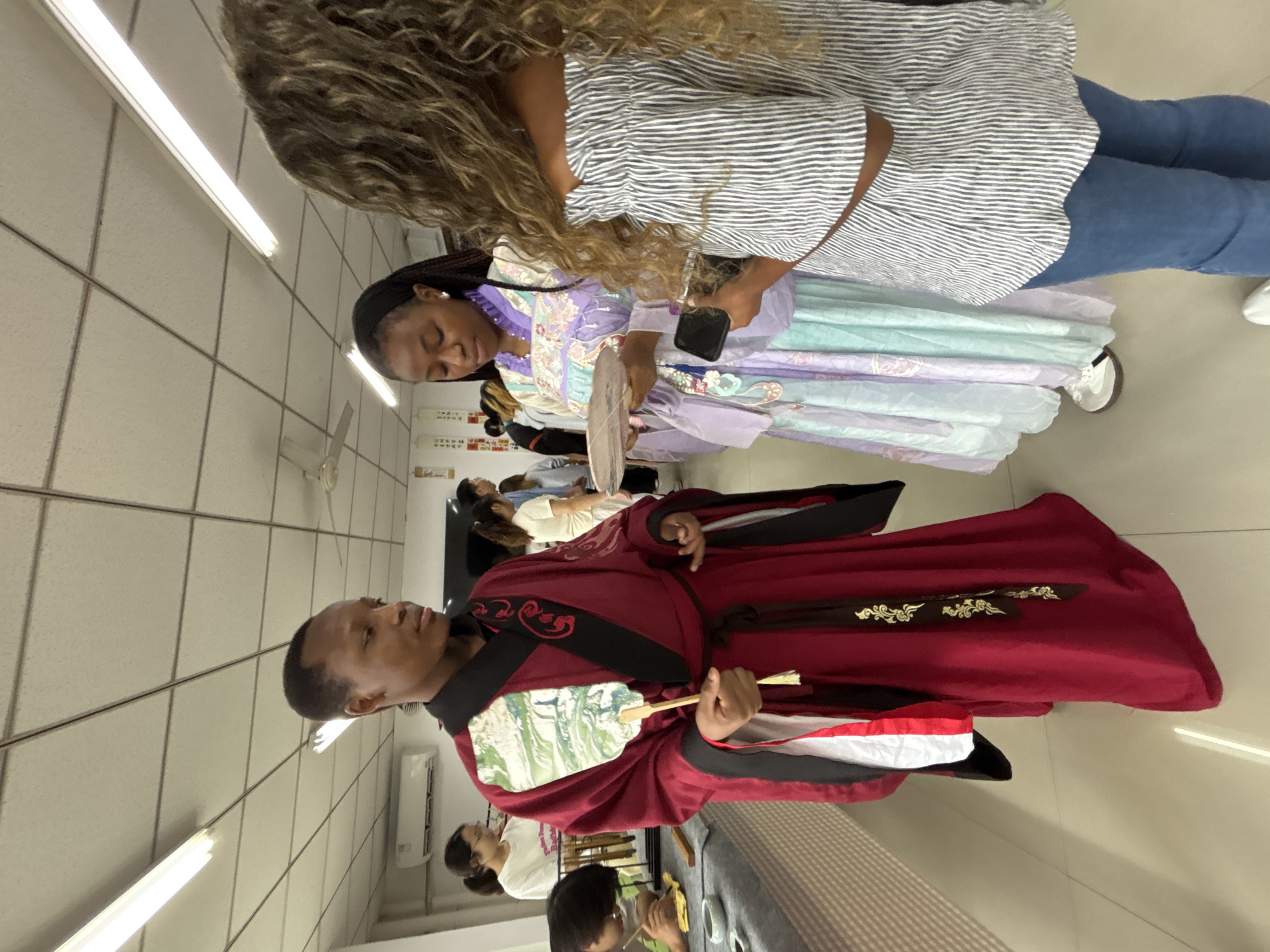 The image depicts three individuals indoors in a brightly lit space with fluorescent ceiling lights. One person is standing in front dressed in traditional Chinese clothing, specifically a red Hanfu robe with black and gold detailing and holding a fan. Another person standing near them is dressed in a lilac-colored Hanfu with mint-green accents. They are holding a mobile phone and also holding a fan. The third person standing is wearing blue jeans and a striped button-down shirt. Other people are seen seated in the background, some engaging in what appears to be a tea ceremony. The image was taken in the city of Jiangjin in China.