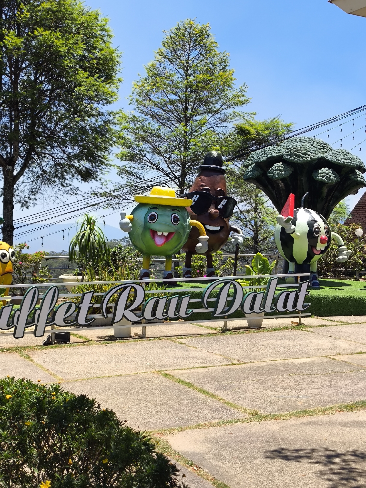 The image shows an outdoor scene in what is likely a park or landscaped area. In the foreground, there is a sign that reads "Buffet Rau-Dalat." Behind the sign, there are several cartoonish statues of produce including a green fruit/vegetable wearing a yellow hat, a potato-like figure wearing sunglasses and a hat, and a watermelon holding a slice of watermelon. These statues are located on a raised platform covered in artificial green grass.
The background includes trees, shrubbery, and possibly some mountainous terrain in the distance under a bright blue sky. The scene is well-lit, suggesting it's daytime. Above, several electrical lines are visible amongst the trees. The overall impression is that this is a tourist attraction or a whimsical advertisement for a restaurant or buffet in Da Lat, Vietnam, specializing in vegetables ("Rau" means vegetable in Vietnamese). The ground in the foreground is made of concrete slabs. A bush with small yellow flowers is on the bottom left.