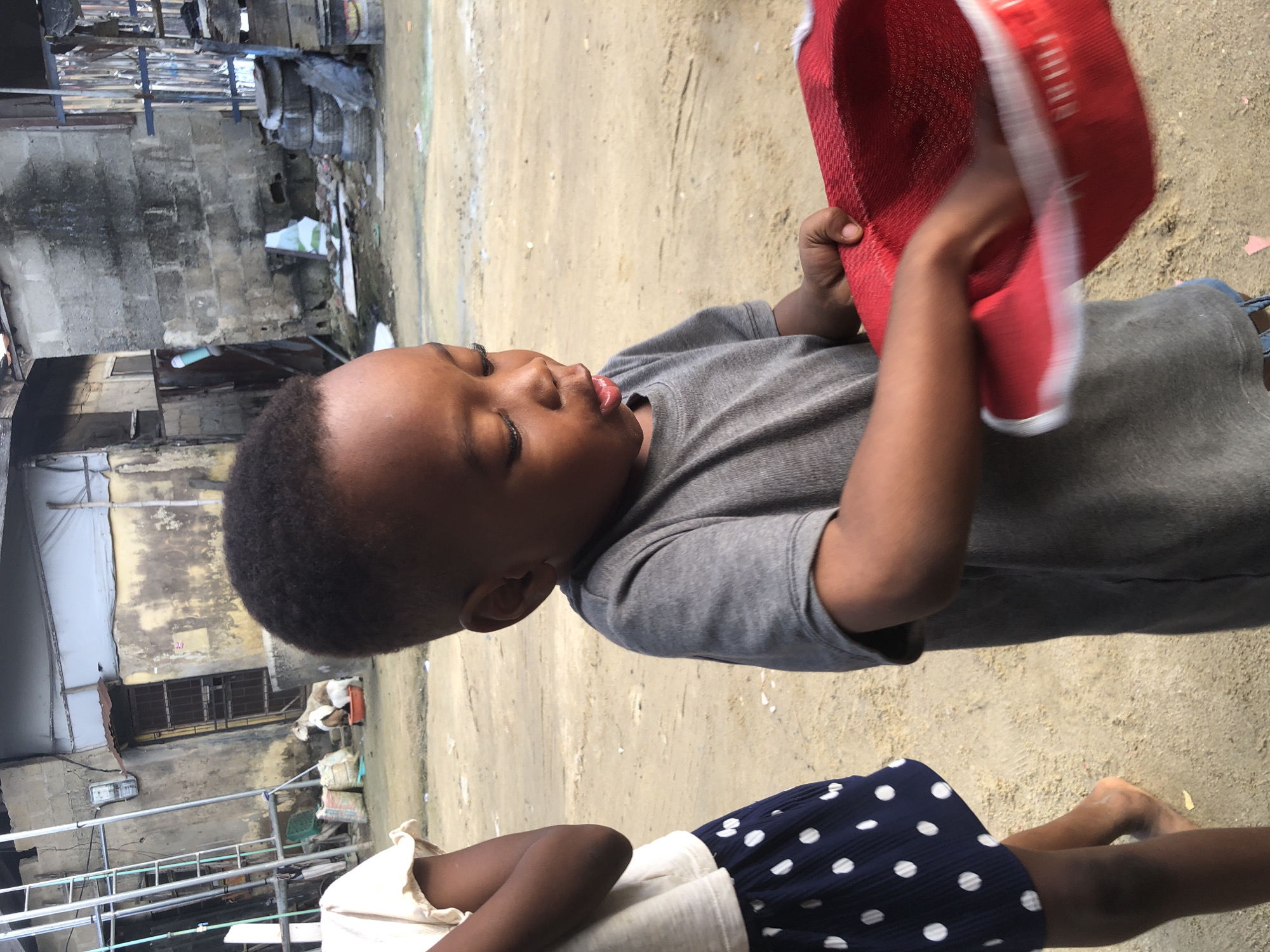 In Ado, Nigeria, a young Black boy, appearing to be between two and three years old, is the central subject of this image. He is wearing a gray t-shirt and blue denim shorts. His attention is focused downwards on a bright red woven hat with a white trim, which he is holding with both hands. His mouth is slightly open, and his expression suggests concentration or curiosity. To the left of the boy, another child, whose face is not visible, is partially in the frame. This child is wearing a light-colored ruffled shirt and a dark blue skirt with white polka dots, and is barefoot.
The environment is an outdoor, dusty area, likely a street or an open lot within a densely populated urban setting. In the background, there are the facades of buildings constructed from concrete blocks and corrugated metal. One building has a yellow-stained exterior and a metal-barred door. To the left, a tall, industrial-looking metal ladder stands against a wall. Various objects are scattered around, including bags of what appear to be supplies, discarded tires stacked up, and possibly some refuse. The overall impression is of a less-developed, possibly impoverished urban neighborhood.