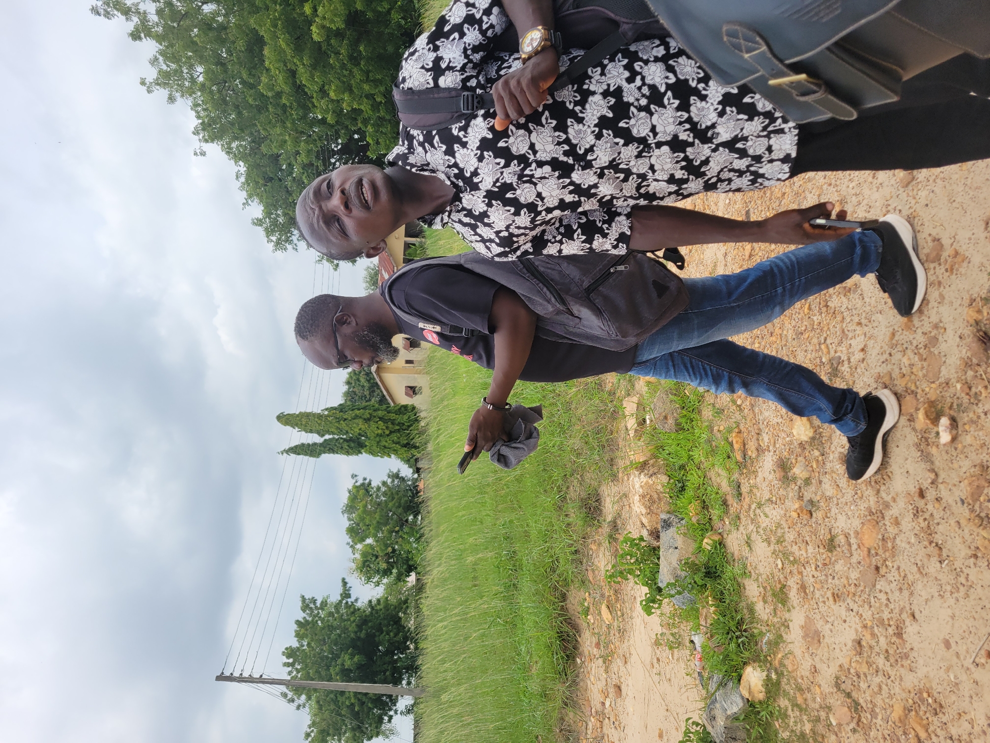 The image depicts two men walking on a dirt path in Keffi, Nigeria. The man in the foreground is wearing a black shirt with a white floral pattern, a backpack, and a watch. He is looking upwards and to the right. The second man, to his left, is wearing a black t-shirt, blue jeans, and sneakers. He is holding a dark cloth and a smartphone in his hands and looking down at the phone. The environment features tall green grass, scattered rocks, and some trees in the background, with a building and utility poles visible in the distance under an overcast sky.