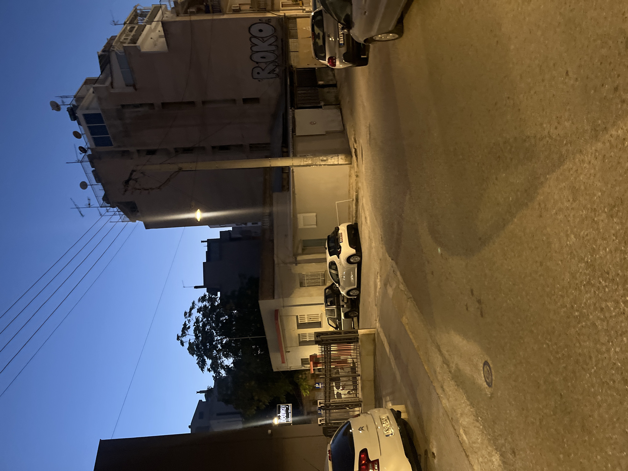 The image captures an outdoor urban scene in Athens, Greece, during twilight. In the foreground, a paved street is visible with several parked cars, including a white Citroën to the left and a silver car further down the street. To the left of the street, a black metal fence encloses a small area with some vehicles, and a sign for "Bedway Athens Hostel" is illuminated. Behind the fence, a low-rise building with windows and a red-trimmed awning is present. Further back, a dark-colored SUV is parked near a building. Dominating the background is a multi-story building with numerous satellite dishes and antennas on its roof. Graffiti that reads "ROKO" is spray-painted on the side of this building. The sky is a deep blue, characteristic of dusk, with power lines stretching across the upper portion of the image. Trees are visible behind the lower buildings, silhouetted against the sky.