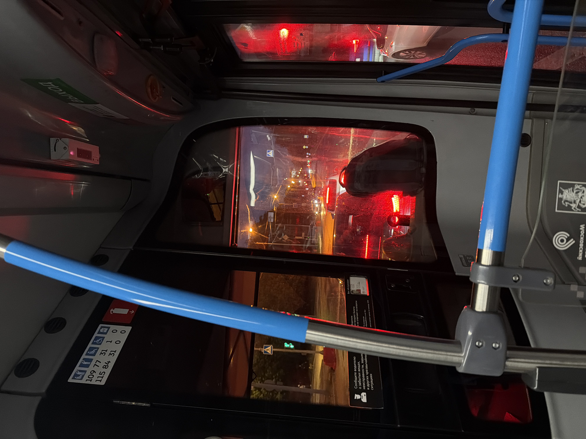 The image shows the interior of a bus at night, looking out through the front windshield and side windows. The scene is illuminated by the red taillights of traffic and the yellow glow of streetlights. In the foreground, a blue handrail is prominently displayed. The bus appears to be stopped in traffic. Signs with Russian text are visible on the interior of the bus. On the exterior, traffic and streetlights are visible. The overall impression is of a typical urban nighttime scene experienced from within a public transportation vehicle.