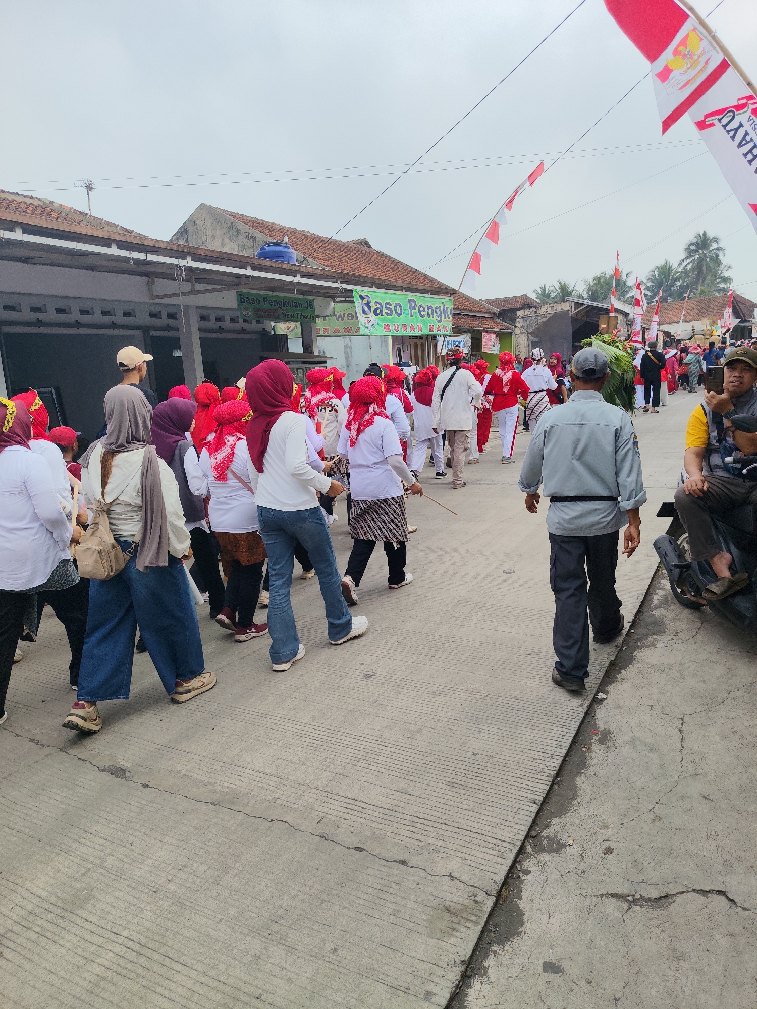 The image depicts a procession or parade taking place on a street in Sukaratu, Indonesia. The street appears to be paved with concrete, and there are buildings with tiled roofs on either side. The sky is overcast. Many people are participating in the event, most of whom are wearing red and white attire, including headscarves and shirts, possibly in celebration of Indonesian Independence Day, as suggested by the presence of numerous Indonesian flags and red and white banners.
In the foreground, several individuals are walking. On the left, a person in a white shirt, blue jeans, and a beige backpack is walking away from the camera. Next to them, another person in a white shirt and blue jeans, with a red headscarf, is also walking forward. To the right of this group, a man in a grey shirt and dark pants, wearing a baseball cap, is walking away from the camera. He appears to be in a more formal or organized role, possibly security or event staff. In the mid-ground, a larger group of people in matching red and white outfits are moving along the street, many with red headscarves. Some are walking in pairs or small groups, while others are in a more cohesive line.
On the right side of the image, a man is sitting on a motorcycle, holding a mobile phone up as if to take a picture or record a video. He is wearing a yellow shirt and dark pants. Other people are visible in the background, participating in the event or observing from the sides. Flags, both large Indonesian flags and smaller red and white banners, are prominent throughout the scene, indicating a festive or patriotic occasion. Signs with text, though largely unreadable due to the distance and resolution, are also visible attached to the buildings. The overall atmosphere appears to be lively and communal.