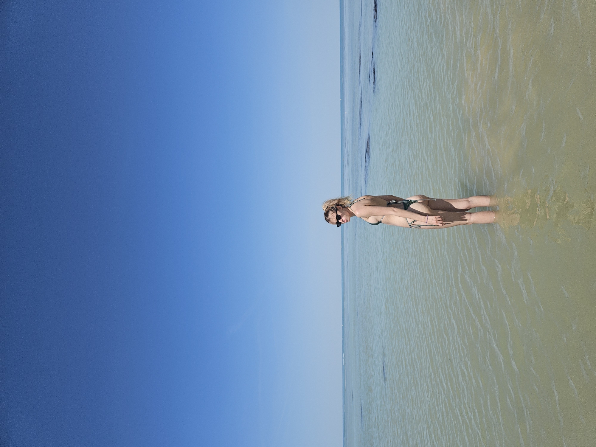 A young woman stands waist-deep in shallow water at a beach in La Brée-les-Bains, France. She is wearing sunglasses and a bikini. The water stretches out to the horizon, meeting a clear blue sky. There are some darker patches in the water, possibly seaweed. The scene is brightly lit.