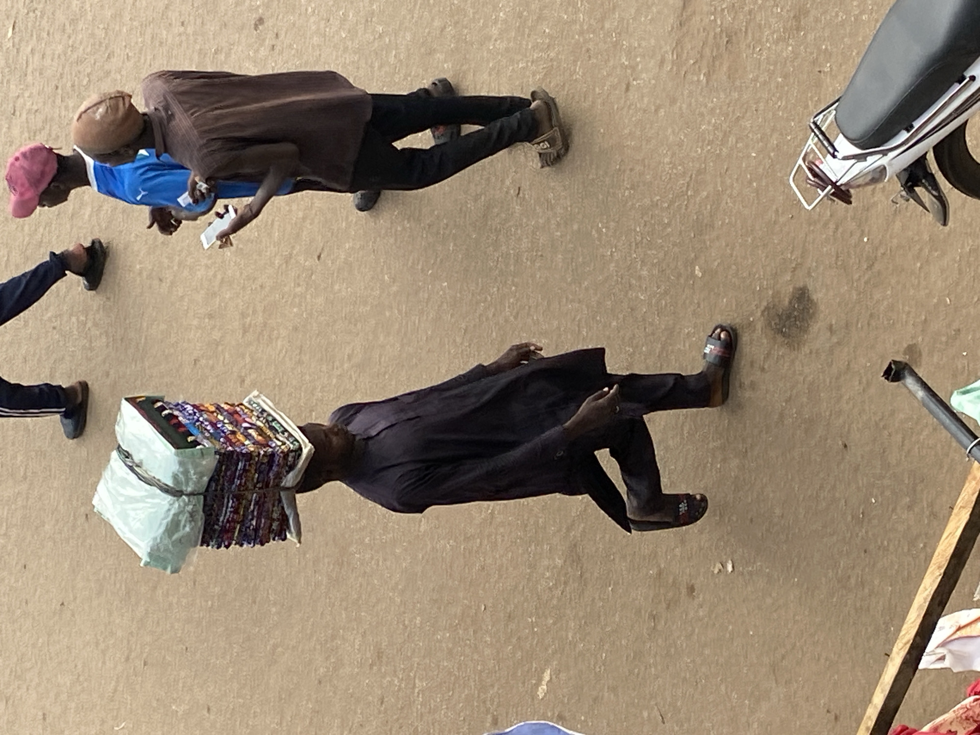The scene depicts a street or open market area in Bukuru, Nigeria, observed from an elevated perspective. The ground is unpaved, consisting of light brown earth with minor darker smudges.
In the left-center foreground, a dark-skinned man walks from left to right across the frame. He wears a dark, long-sleeved tunic (possibly a *kaftan*) and dark trousers, with black open-toed slides on his feet. He balances a tall, rectangular stack of merchandise on a flat, light-colored tray or board on his head. The lower portion of the stack reveals multiple layers of folded, colorful textiles or fabrics. The upper portion is wrapped in a light green plastic bag, secured with dark straps.
Towards the right side of the frame, two other dark-skinned men are engaged in an interaction. The man on the far right wears a brown long-sleeved button-up shirt, dark trousers, and light brown open-toed sandals. A light brown head covering, possibly a cap or wrap, is on his head. He holds a white smartphone in his left hand, appearing to display or examine it. Facing him, the other man wears a blue short-sleeved t-shirt (resembling a sports jersey), dark trousers, and dark footwear. He has a pink baseball cap on his head and his hands are partially raised in front of him, facing the man with the smartphone.
In the far background to the left, only the lower legs of another person are visible, wearing dark trousers with a white stripe and dark sandals. The rear section of a white motorcycle or scooter, featuring a black seat and a silver metal rack, is visible in the bottom right corner of the frame. In the bottom left, a wooden beam or plank with some white and red fabric draped over it is partially visible.