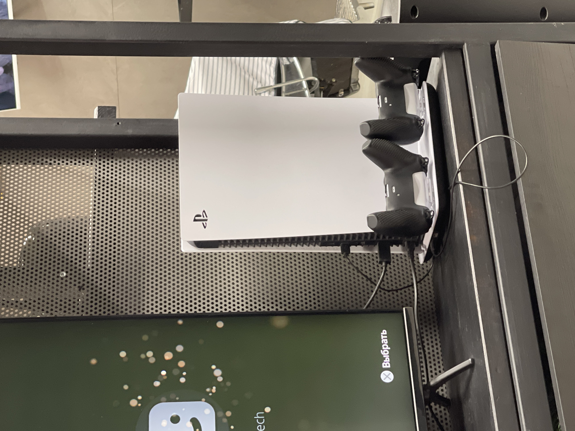 The image shows a PlayStation 5 console with two controllers mounted on a dark grey perforated metal shelf. Below the shelf, a portion of a screen is visible, displaying text in Russian (Выбрать, "Select"). The scene appears to be indoors, within a black metal frame structure. It is impossible to determine the location specifically in Saint Petersburg, Russia without further context or identifying features.