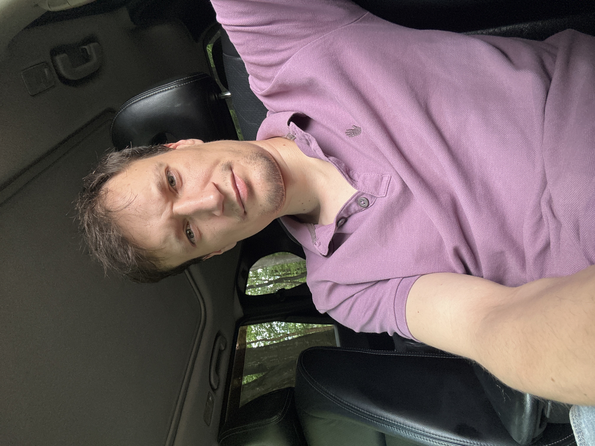 A man with light brown hair and a light complexion is seated in the driver's seat of a vehicle. He is wearing a purple polo shirt. The interior of the car appears to be black leather. Through the car windows, trees and foliage are visible, suggesting an outdoor, possibly wooded, location. The image appears to be a selfie taken inside the car. The specific location in Shatava, Ukraine, is not identifiable from the visual cues in the image.