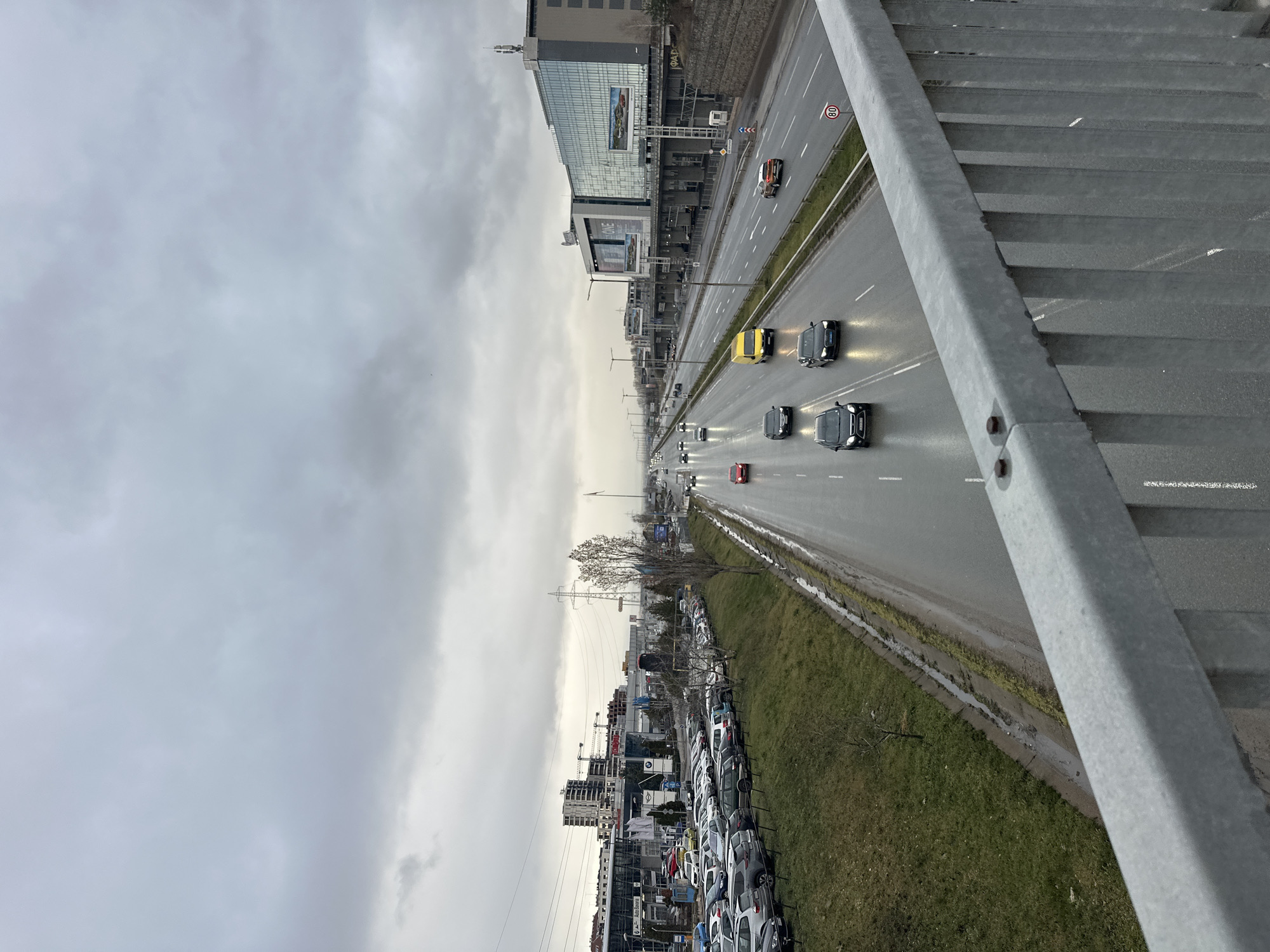 The image depicts a view from a pedestrian overpass in Sofia, Bulgaria. Below the overpass is a multi-lane highway with vehicles traveling in both directions. The road surface appears wet, suggesting recent rain. On one side of the highway is a row of parked cars, likely for sale, with several commercial buildings in the background, including a dealership. On the opposite side of the highway is a large commercial building with glass and steel architecture. The sky is overcast, contributing to the overall grey tones of the scene. The overpass railing is in the foreground, providing a visual frame to the view.