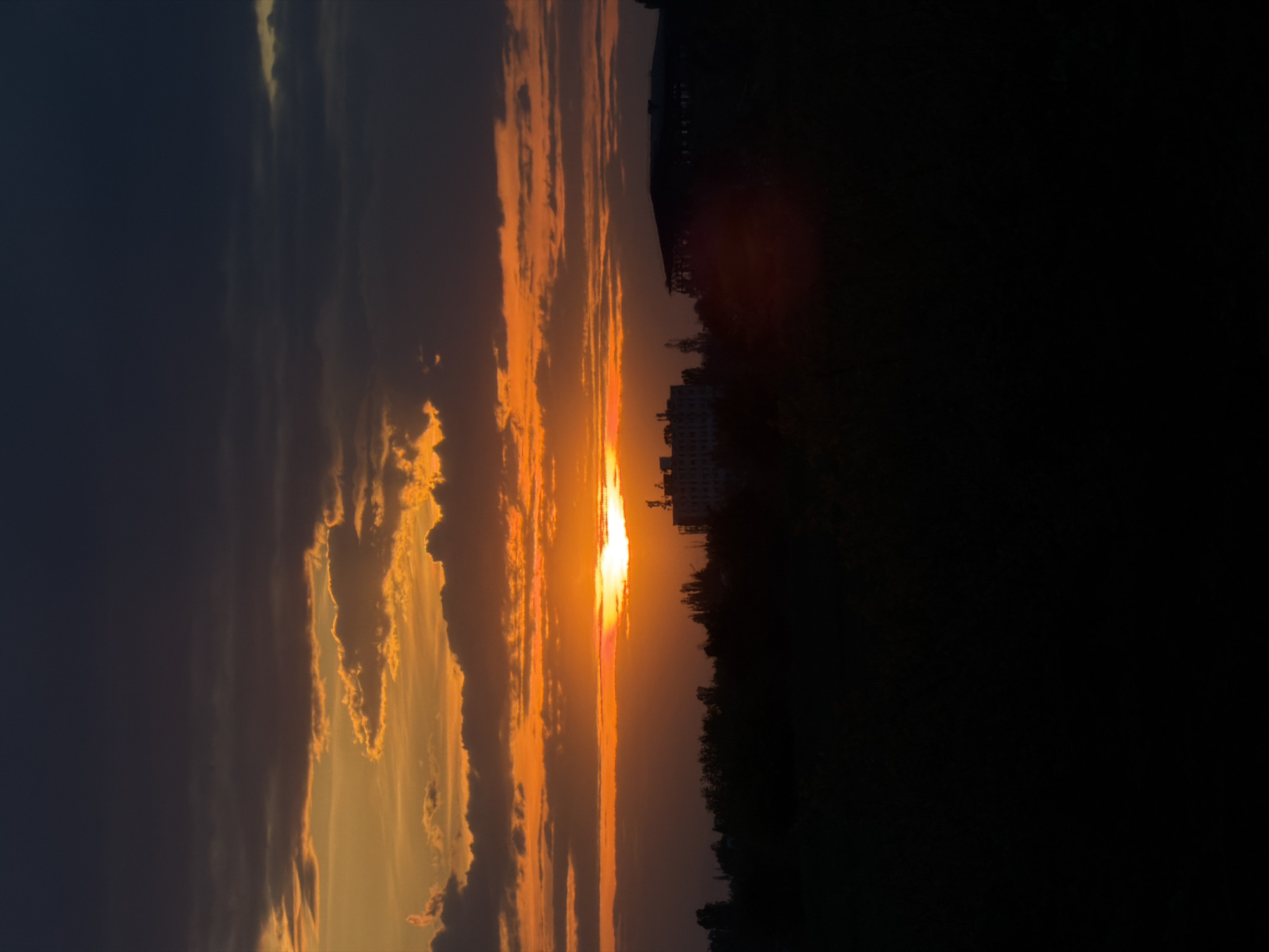 The image depicts a sunset over what appears to be a landscape or cityscape, with the sun positioned centrally between dark, silhouetted forms on the right and colorful, illuminated clouds on the left. The sky is streaked with orange and yellow hues, indicative of a setting sun. A faint silhouette of buildings is visible on the right side of the frame, set against the darker backdrop. The scene suggests a location with some elevation, as the view encompasses both the sky and distant features. It is an outdoor shot, capturing a moment in time with natural lighting conditions. Without further details, the location in Yana, Bulgaria cannot be confirmed.