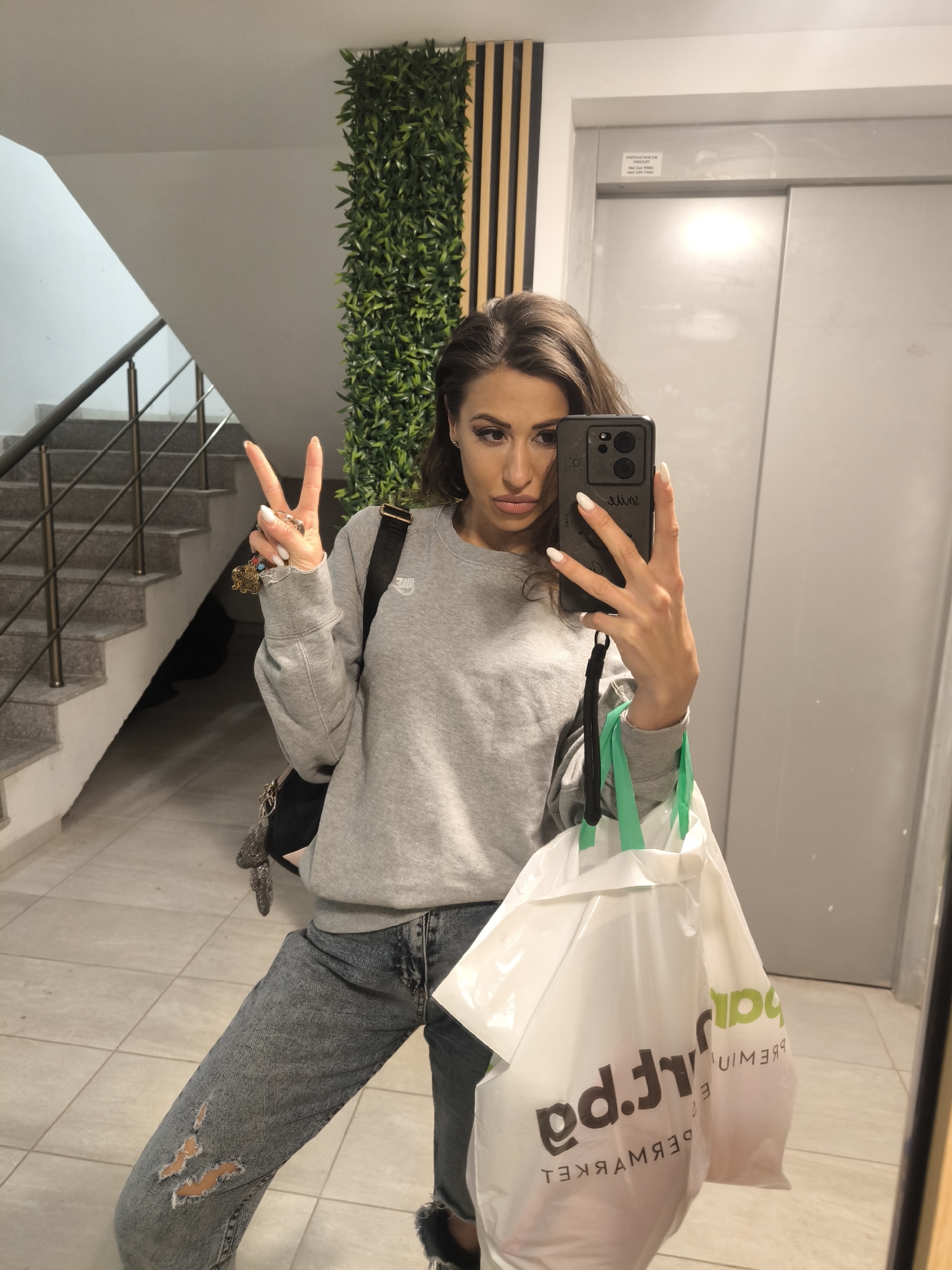 A woman stands indoors, likely within an apartment building, as indicated by the presence of a staircase on the left and an elevator door behind her. She holds a smartphone in her right hand, presumably taking a selfie, and makes a peace sign with her left hand. She's wearing a gray sweatshirt and ripped jeans and a backpack. She carries white shopping bags with green handles, identifiable by the reversed words "market premium" and the letters "pa" or "ba". Behind her is an ornamental green wall, along with a contrasting wooden slat design. The flooring appears to be light-colored tiles. The city is unknown.
