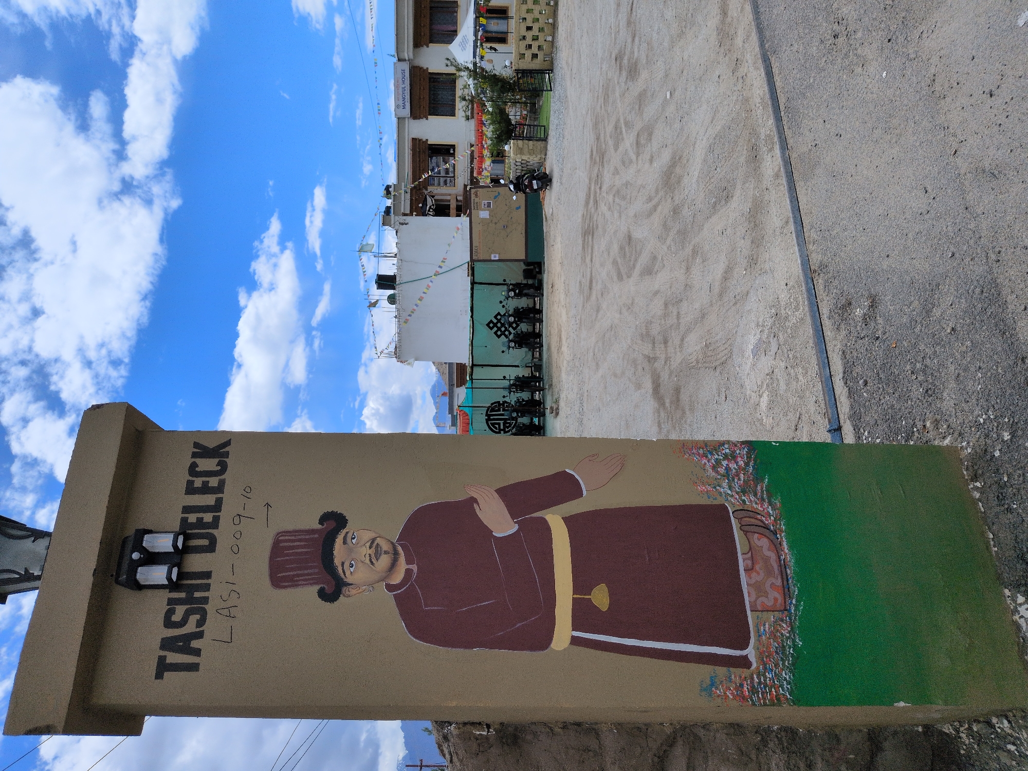 In the city of Leh, India, a tall, tan-colored pillar stands at the foreground, adorned with a painted portrait of a man wearing a maroon hat and traditional attire. The man has dark hair, a mustache, and is depicted with his arms outstretched. Text above the portrait reads "TASHI DELECK" and "LASI-009-10." The pillar is illuminated by two black lanterns mounted at the top.
Behind the pillar, a gravel courtyard is visible, with several motorcycles parked near a building. The building, identified by a sign as "MANGYUL HOUSE," is a white, two-story structure with dark windows and wooden accents. Colorful prayer flags are strung across the front of the building and on its roof. Further in the background, mountains can be seen against a bright blue sky dotted with white clouds. The scene captures an outdoor setting, likely a guesthouse or local establishment, in the Himalayan region.