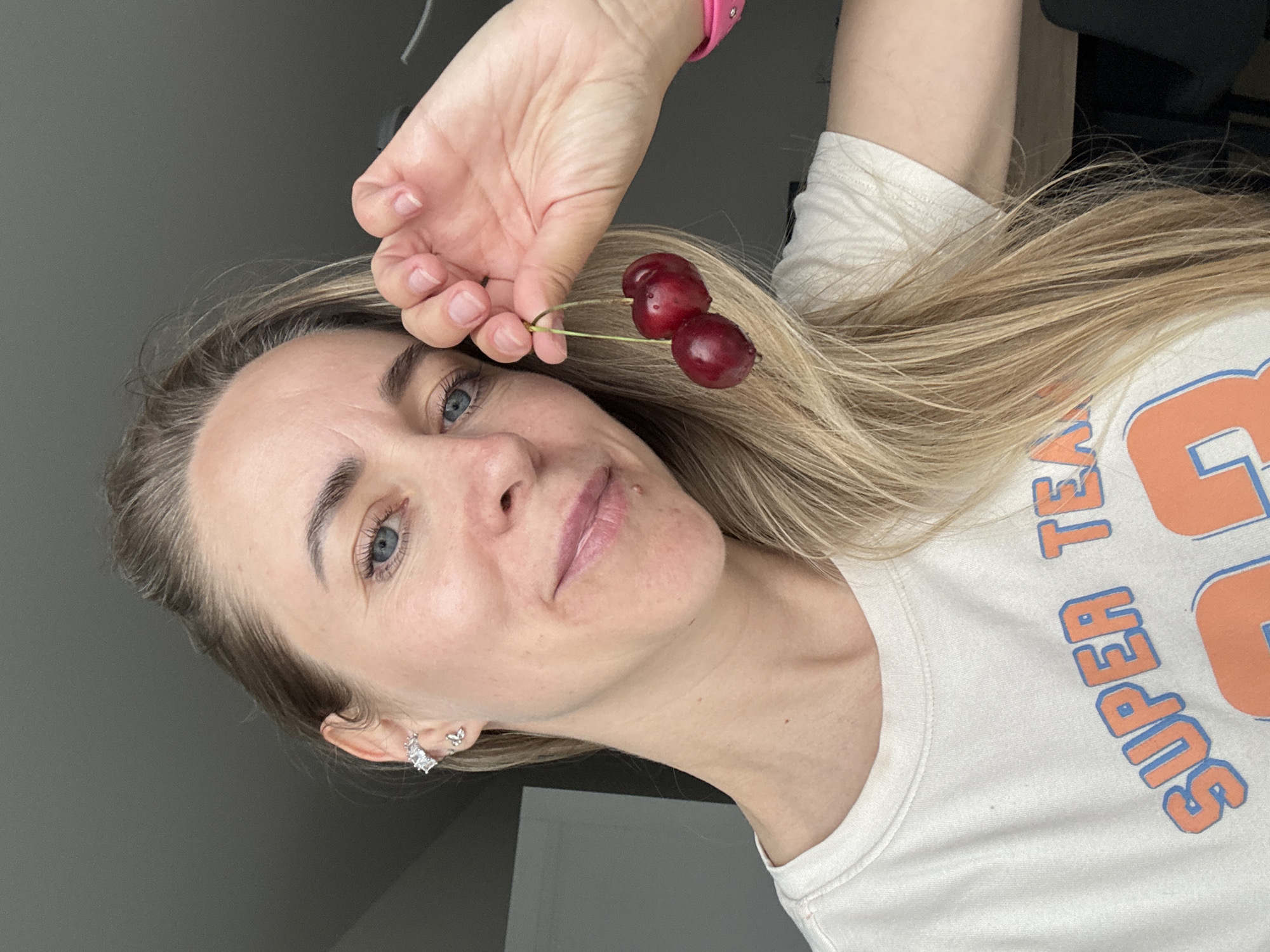 An eye-level, portrait-style shot captures a woman indoors, her head and upper torso angled, suggesting the image is rotated on its side. She has fair skin, light blonde hair, and blue eyes, with a calm, subtle expression and a faint smile. Her left ear features two silver earrings: a rectangular hoop and a smaller, possibly butterfly-shaped stud.
In her right hand, she holds a pair of dark red cherries by their green stems, positioning them near her hair on the right side of the frame. A pink wristband is visible on her right wrist. She wears a light-colored t-shirt with bold orange letters outlined in blue, partially reading "SUPER TE". The background is a plain, dark grey wall, indicating an indoor setting with diffused lighting. The overall mood is relaxed and casual. The provided context indicates the location is Chaikovsky, Russia.