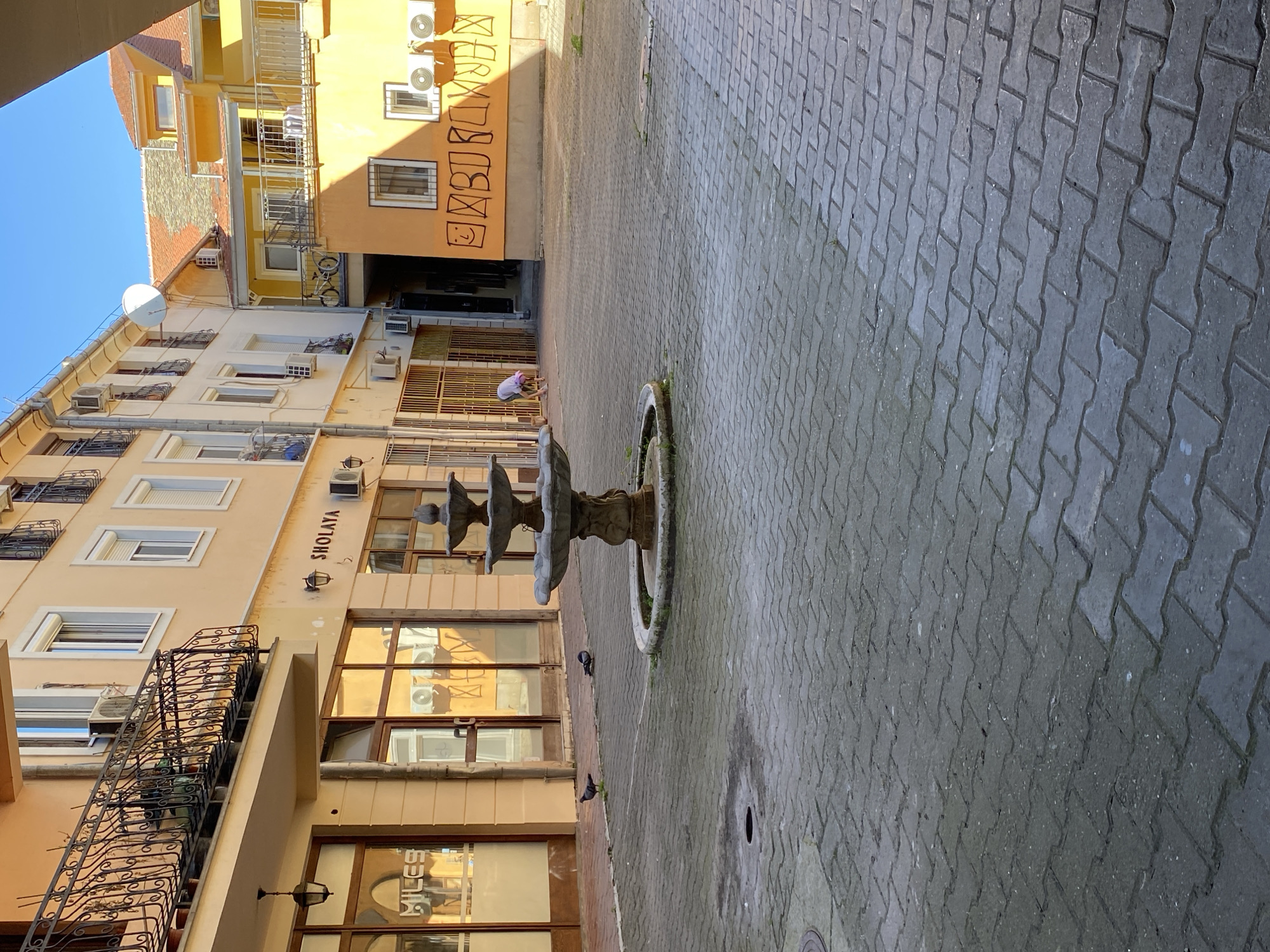 A cobblestone courtyard in Novi Sad, Serbia, features a stone fountain with three tiers and a circular basin. Several pigeons are present near the fountain. The courtyard is surrounded by buildings, one with a sign reading "SHOLAYA." Graffiti is visible on the upper portion of a building. A person is bent over near an open gate. The sky is partially visible, a clear blue.