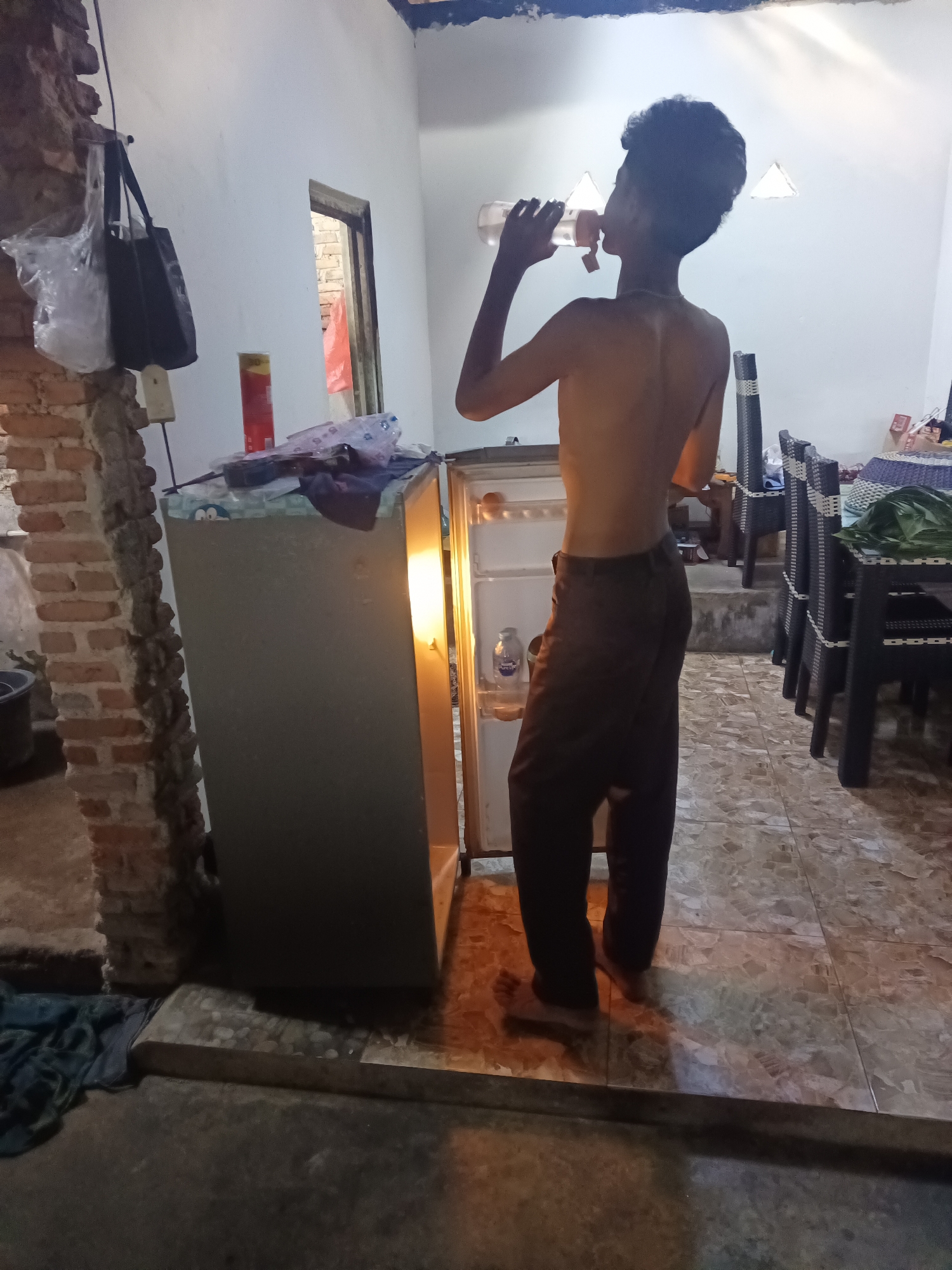 A shirtless young male with dark curly hair stands in front of an open, illuminated refrigerator, drinking from a clear bottle. He is wearing dark-colored pants. The refrigerator is positioned in a room with white walls and a tiled floor. To the left of the refrigerator, a rough brick wall is visible, with a black handbag and a plastic bag hanging from a hook. On top of the refrigerator are various items, including a red and yellow can and some plastic packaging. In the background, there are chairs and tables. The scene appears to be indoors, likely a residential setting. The location provided, Terbanggi Marga, Indonesia, suggests a tropical or subtropical climate, which is consistent with the indoor setting and the type of building materials shown.