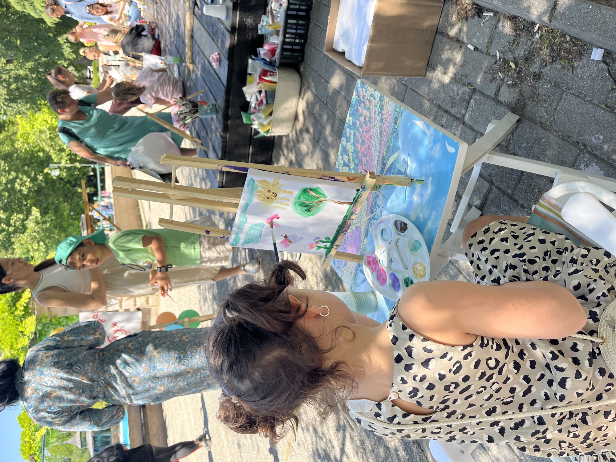 A medium shot presents a group of people engaged in an outdoor painting session in a public space on what appears to be a sunny day. Focus is given to a young woman with brown hair pulled up in two buns. She wears a beige and black patterned dress and is seated at a painting easel where she is working on a painting of a tree, sun, and clouds. A palette of paints is visible beside her. In the background, other people are also painting at easels, and several individuals are seated on a low wooden platform. Baskets and boxes of art supplies are scattered around. The setting is likely a park or public area where an art event is taking place. Although the location within Plovdiv, Bulgaria, cannot be precisely determined from the image alone, the presence of people engaged in an art activity in a public outdoor space is clear.