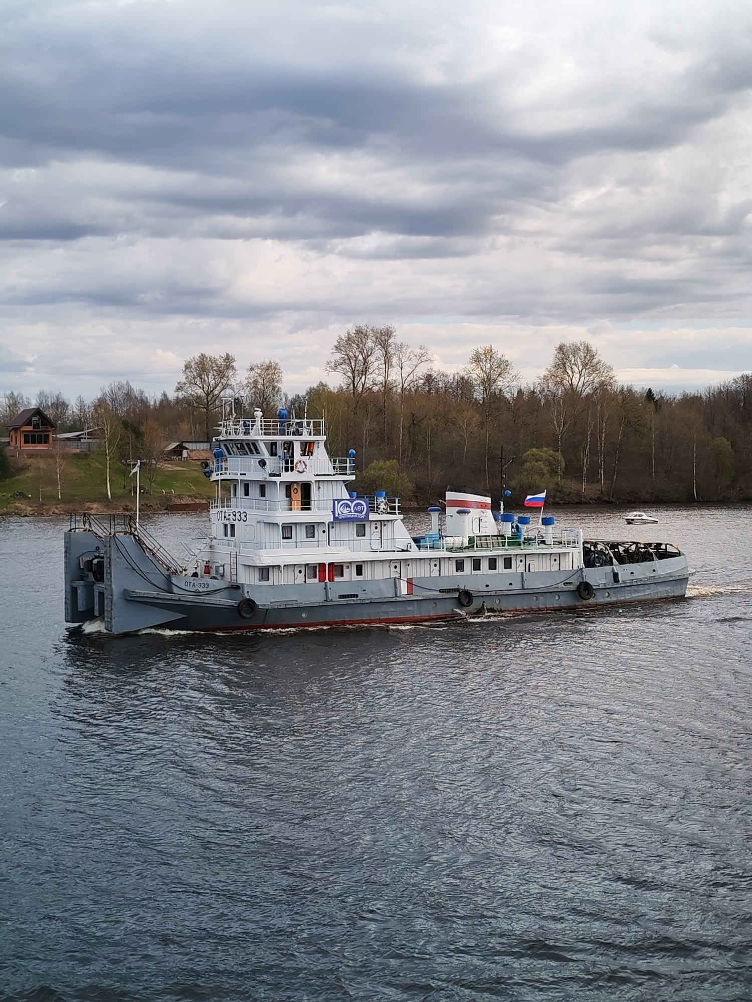 The image depicts a gray tugboat traveling on a body of water, likely a river or a wide canal, in what appears to be the vicinity of Novoe Selo, Russia. The vessel has a multi-tiered superstructure, with the name "OTA-933" visible on its side. A Russian flag is displayed on the stern of the boat. The water is dark and rippled, suggesting a slight breeze. The sky is overcast with heavy gray clouds.
In the background, a forested shoreline with trees that are either bare or just beginning to leaf out can be seen. A few buildings, including a house with a reddish roof, are visible near the riverbank. A small, white motorboat sits still in the water behind the tugboat. The overall scene suggests a rural or suburban setting on a cloudy day.