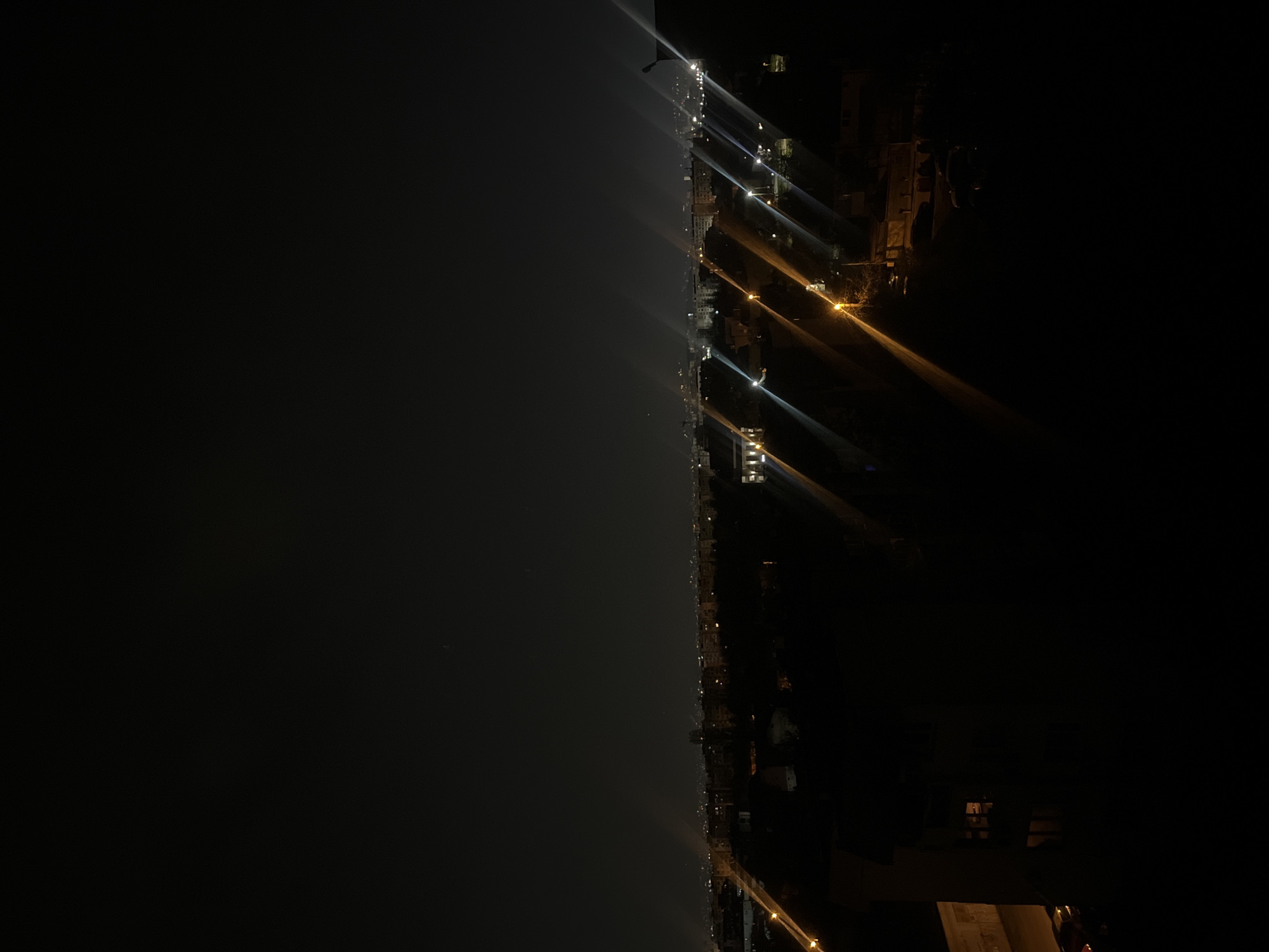 The image depicts a nighttime view of a coastline, possibly a waterfront area within Sofia, Bulgaria. The scene is dominated by darkness, with the left side of the image showing a dark expanse of water or sky. A line of lights extends along the coast, suggesting a road or promenade illuminated by streetlights. The lights cast long beams, possibly due to atmospheric conditions like fog or mist. Buildings are visible on the right side of the image, with some lit windows and architectural details discernible. The photograph appears to have been taken from an elevated position, possibly from a building or higher ground.