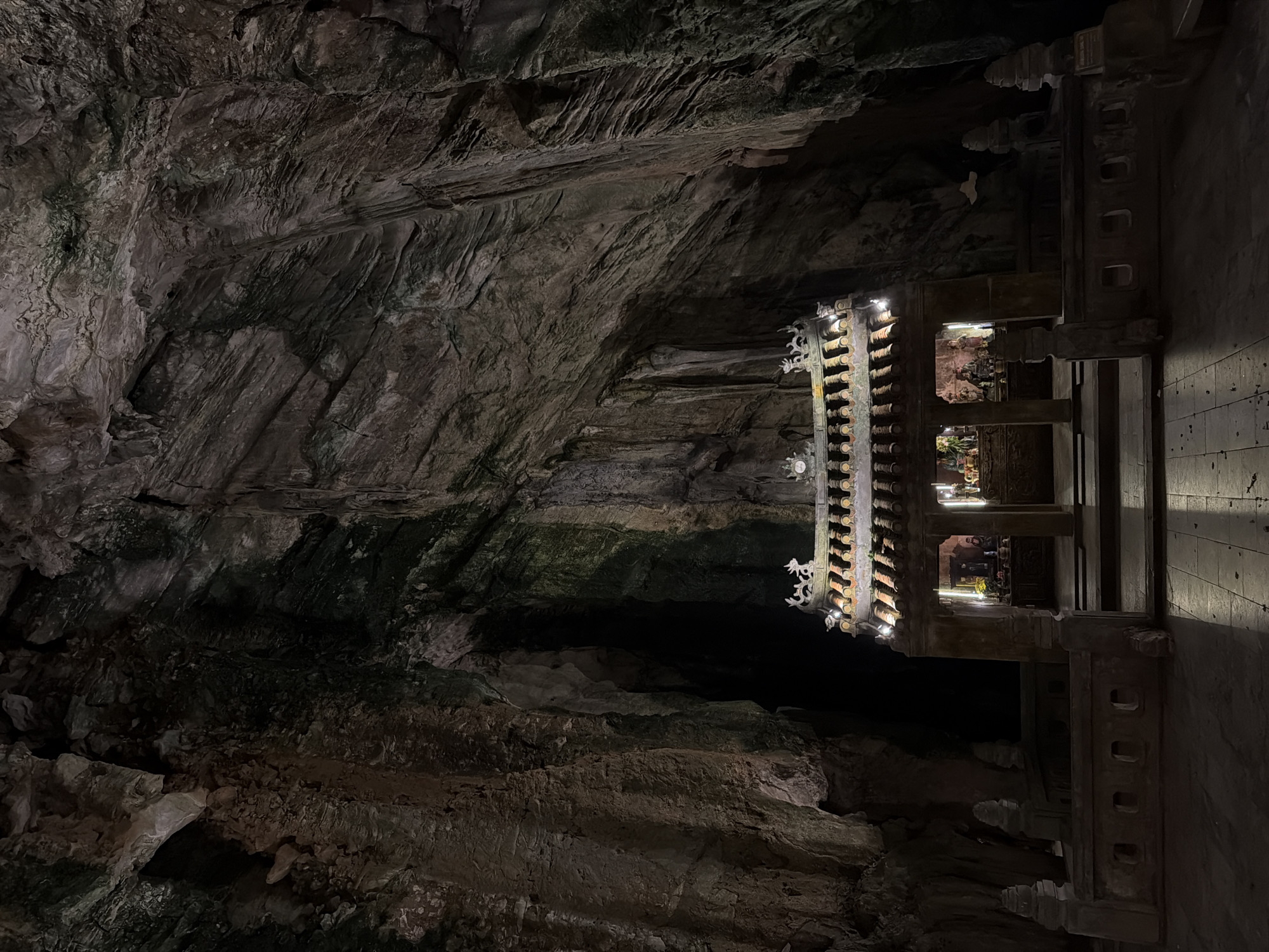 The image depicts a cave interior with a temple structure. The cave walls are rough and dark, with visible rock formations. The temple is constructed from dark-colored materials, possibly wood or stone, with intricate carvings and designs. It is illuminated by multiple small lights, highlighting the details of the structure and revealing religious figures within. The floor around the temple is made of wooden planks. This is likely a view within the Marble Mountains caves in Đà Nẵng, Vietnam.