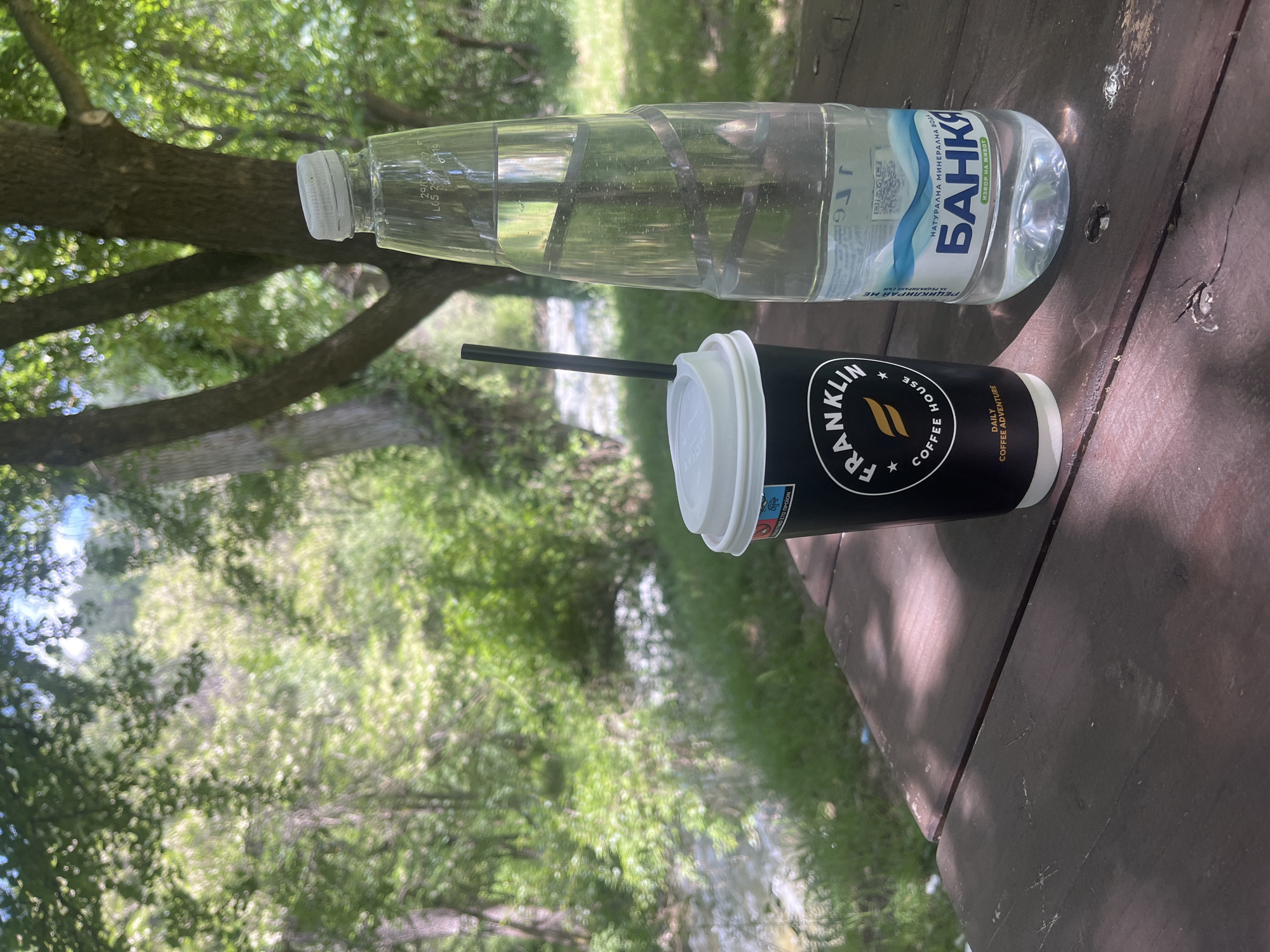 The image shows a bottle of Банкя mineral water and a cup of Franklin House coffee on a wooden table outdoors. The bottle of water is transparent and partially filled, with the Банкя label prominently displayed. The Franklin House coffee cup is black with a white lid and straw, featuring the brand's logo and the text "DAILY COFFEE ADVENTURE." The table is weathered and shows signs of use. The background is blurry and shows a mix of green foliage, suggesting a park or natural setting. The presence of Bulgarian text on the water bottle suggests that the location is likely in Bulgaria. The water bottle label says, "НАТУРАЛНА МИНЕРАЛНА ВОДА ИЗВОР НА ЖИВОТ," and the water bottle is labeled, "БАНКЯ." Considering these factors, it is reasonable to infer that the scene might be located in a park or recreational area in Blagoevgrad, Bulgaria, or another location in Bulgaria.