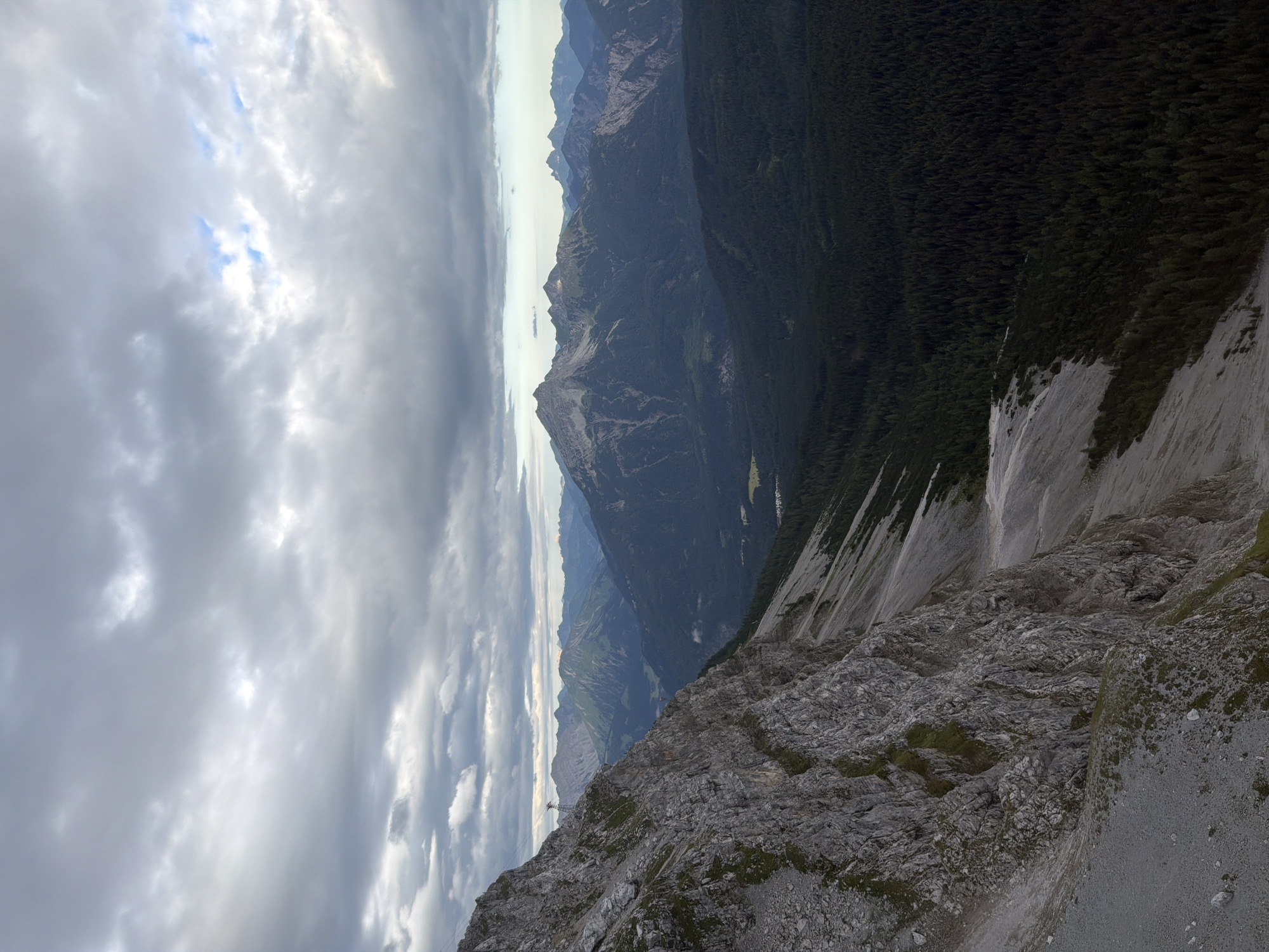 An overcast sky looms over a dramatic mountainous landscape. In the foreground, a steep, rocky mountain face descends from the left side of the frame. The rock is a mix of grey and brown, with patches of sparse green vegetation clinging to its surfaces. A scree slope runs down from the base of the rock face into the mid-ground.
Further into the scene, a dense, dark green coniferous forest covers the valley floor. Beyond the forest, a range of majestic mountains rises, their peaks varying in height and jaggedness. Some of these distant mountains are covered in scree and rock, while others display lush green slopes. The sky is filled with heavy, grey clouds, with occasional breaks revealing hints of blue. A red pylon with cables, suggesting a cable car system, is visible on the left side of the image, near the top of the rocky mountain. The scene depicts a rugged, natural environment, likely in the Bavarian Alps near Grainau, Germany, given the characteristic topography and vegetation. There are no people or animals visible in the image.