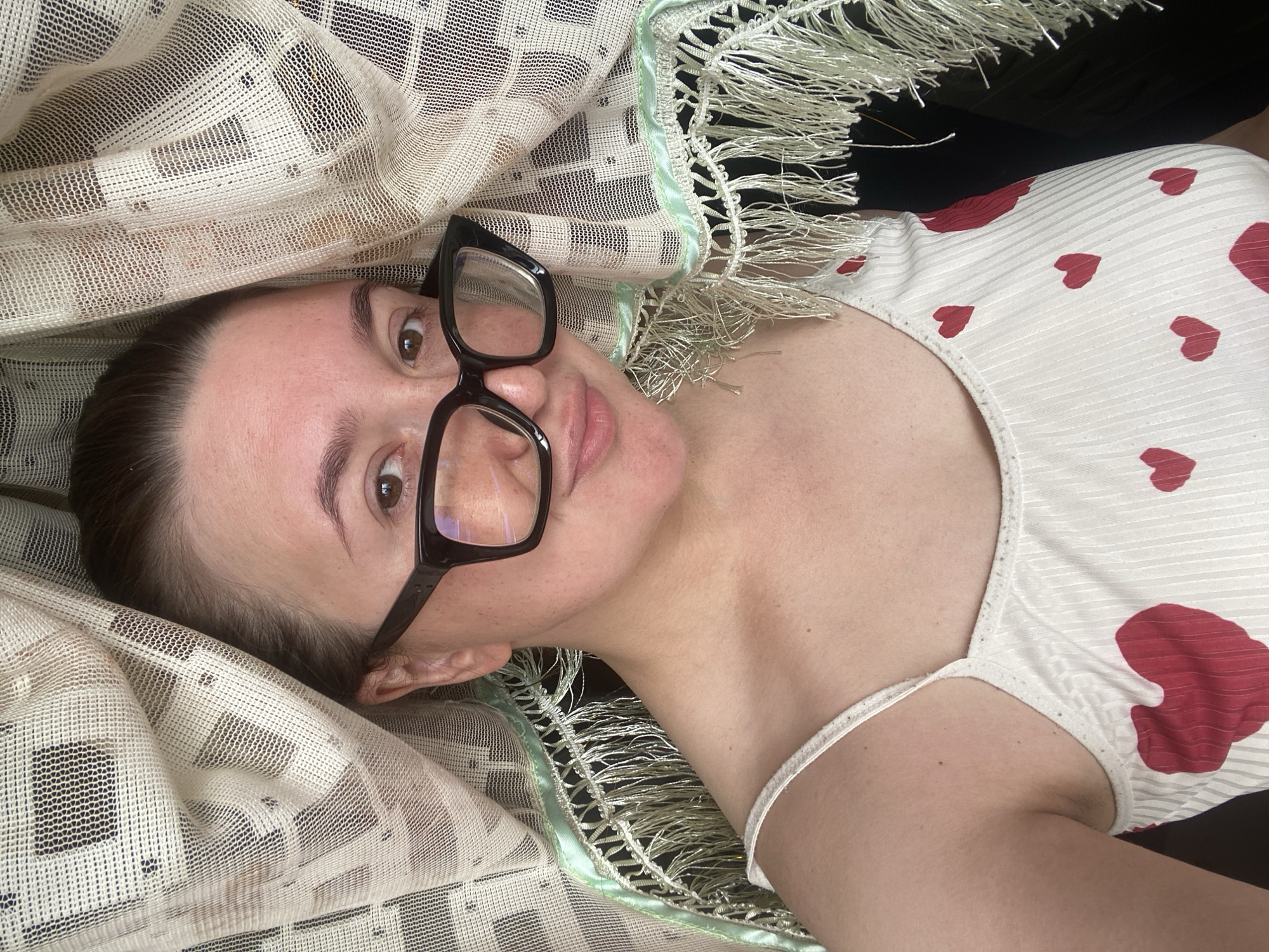 A woman wearing glasses and a white camisole with red hearts is partially obscured by a sheer, patterned curtain with fringe. The woman is looking at the camera and smiling slightly. The scene appears to be indoors. There is no indication of the city of Chaikovsky, Russia, in this image.