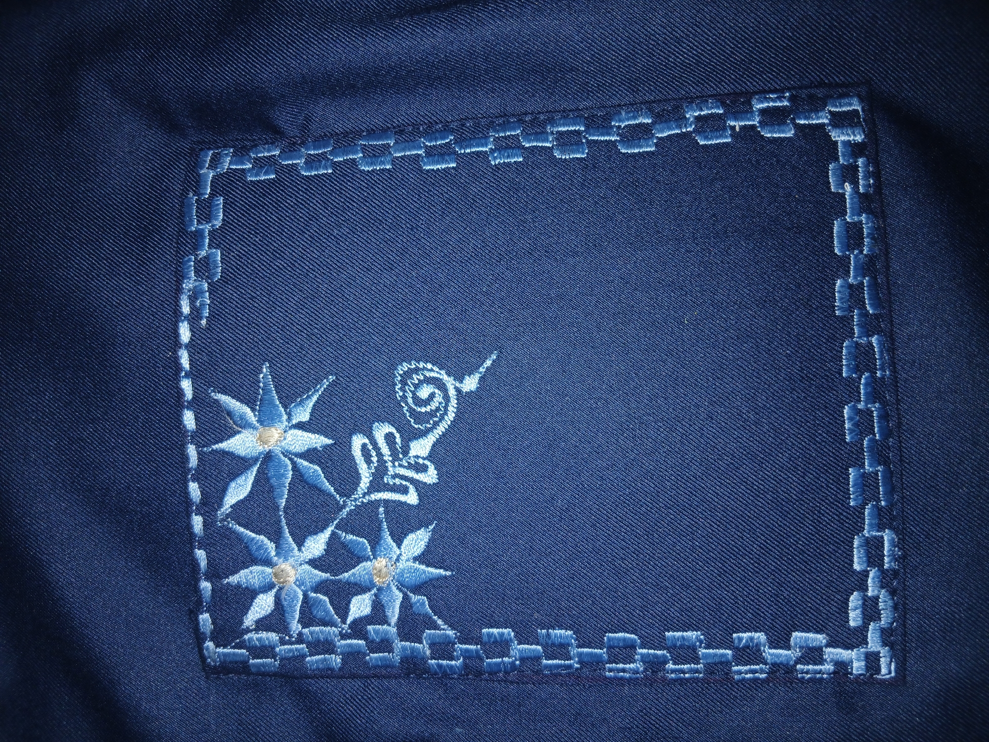 A close-up shot reveals a section of dark blue fabric, possibly part of a garment. The fabric features a rectangular embroidered design in a lighter shade of blue. The embroidery forms a frame around the rectangle with a pattern resembling interconnected blocks. In the lower left corner of the rectangle, there is an embroidered floral design with three stylized flowers and swirling tendrils extending upward. The flowers have pointed petals and a small, round center. The scene does not provide any information to determine the location in Lagos, Nigeria. There are no people, objects, or actions visible in the image that allow me to pinpoint that location.