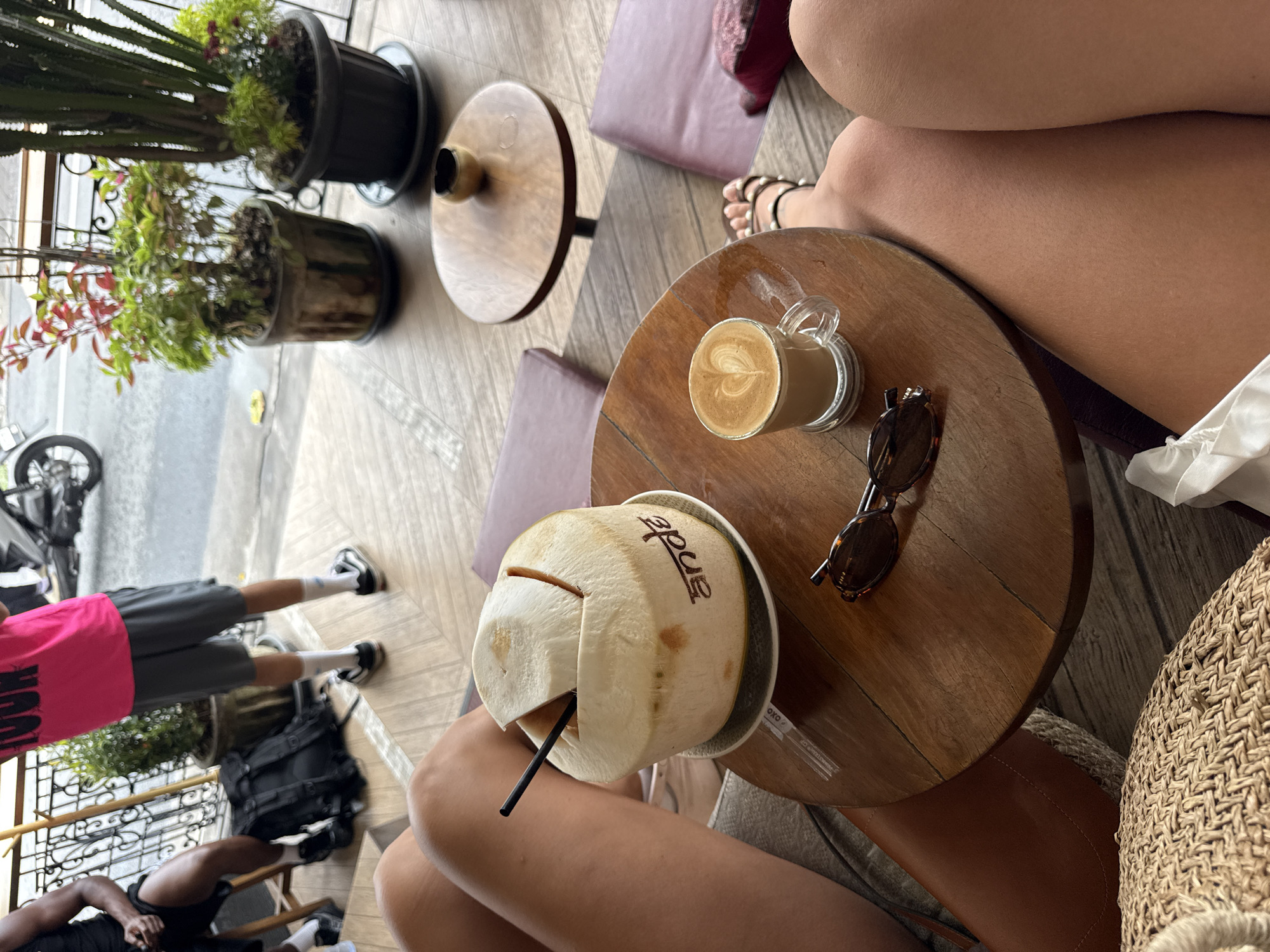 The image depicts a table setting in Kuta Selatan, Indonesia. On the table, there is a latte with latte art in a clear glass mug, a pair of sunglasses, and a coconut with a straw. The coconut has the word "Anda" written on it. Several individuals are seated around the table, their legs visible in the frame. In the background, there are plants in pots, a motorcycle, and a glimpse of the street.