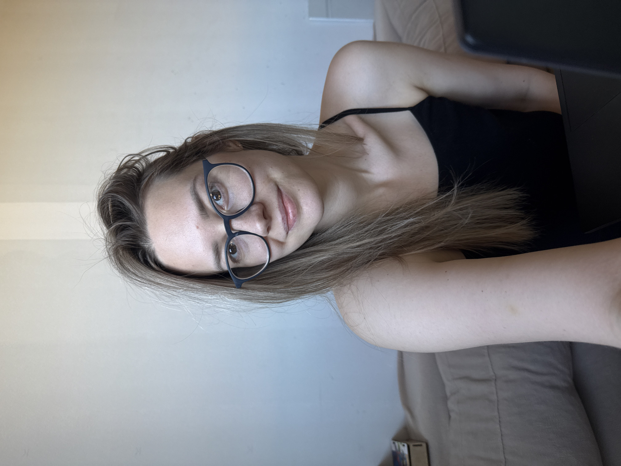 A young woman with light brown hair and glasses is lying on a light brown couch. She is wearing a black tank top. She is looking at a laptop, which is partially visible on the right side of the image. A wall is visible in the background. The location is unknown and cannot be determined to be in Chaikovsky, Russia.