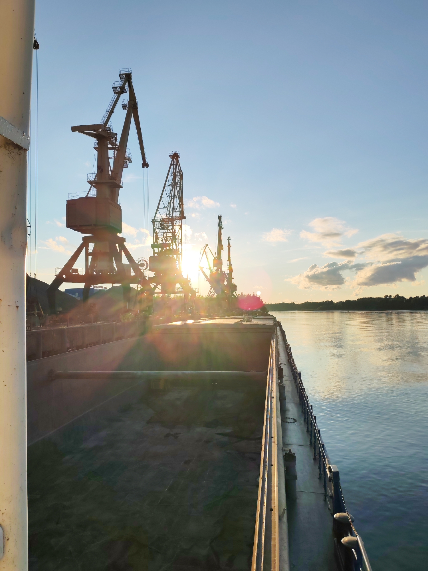 An external, low-angle, wide shot captures a sunrise over a harbor. In the foreground, the deck of a large cargo ship stretches toward the horizon. The ship's open cargo bay is visible on the left, its interior a dark, textured expanse. Along the right edge of the ship, a metal railing runs parallel to the water. The tranquil water of the harbor reflects the warm glow of the sunrise, casting a shimmering light across its surface. In the mid-ground, several large industrial cranes stand silhouetted against the bright sky. The sun, positioned just above the horizon, emits brilliant rays that create a lens flare effect, partially obscuring the cranes. Beyond the cranes, a distant treeline borders the far side of the harbor. The sky is a clear blue, with scattered, wispy clouds tinged by the sunrise.