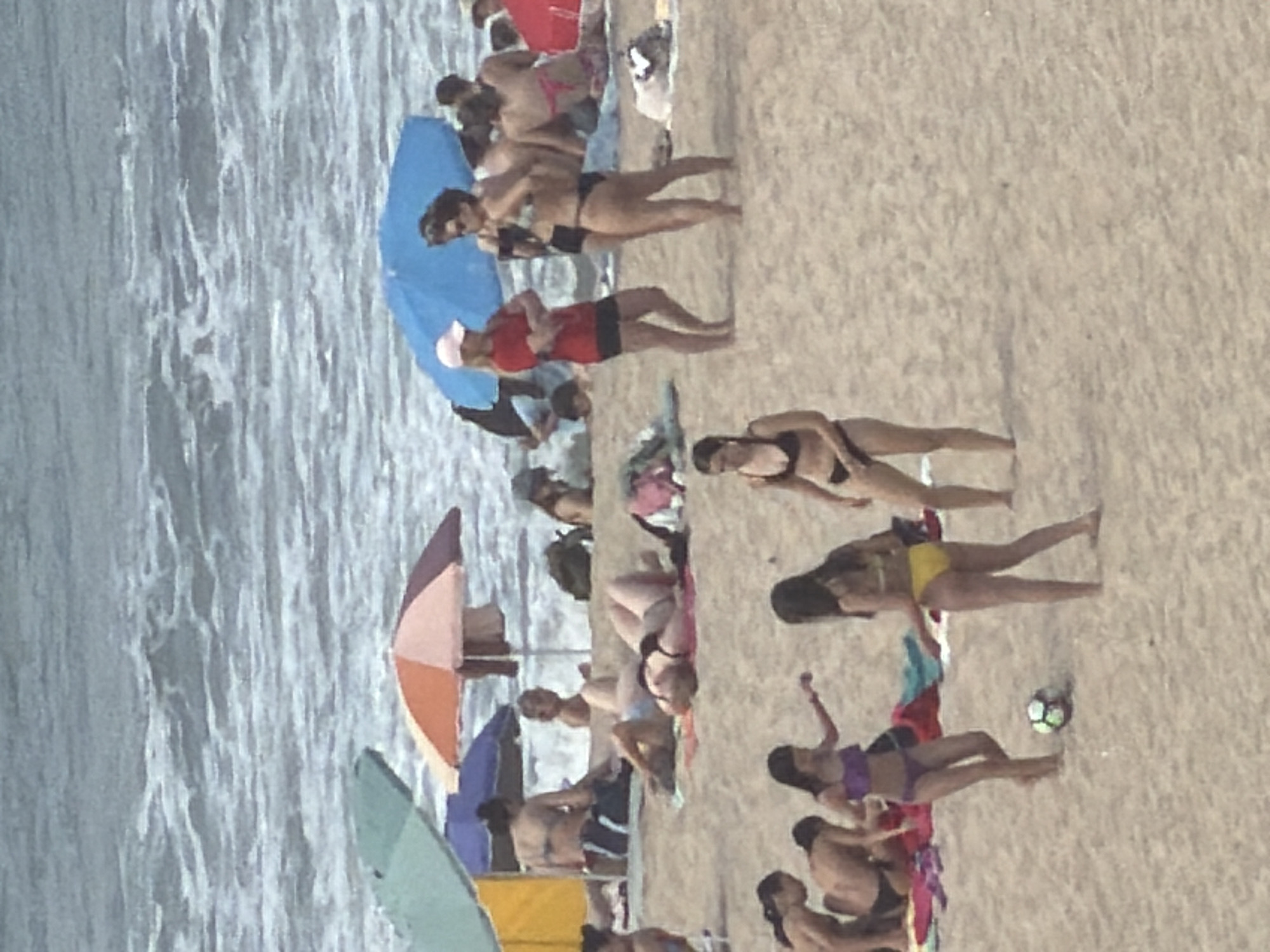 The image captures a crowded beach scene at Praia da Baía in Espinho, Portugal. The ocean is visible in the background with moderate waves. Several beach umbrellas, including a blue one, a green and yellow one, and a red and white striped one, are scattered across the sand. A group of people are engaged in various beach activities. In the foreground, two young women are playing with a soccer ball, one in a purple bikini and the other in a yellow bikini. Other individuals are seen lounging on towels, walking, or standing in groups. The overall atmosphere is one of leisure and recreation on a sunny day.