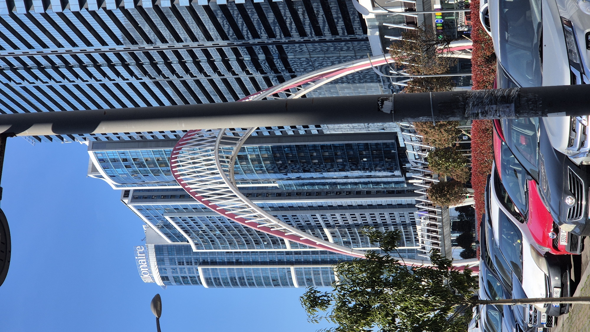The image shows a cityscape in Batumi, Georgia, notable for its modern architecture. The "Billionaire" residential complex stands tall, reflecting the clear blue sky in its glass facade. A pedestrian bridge with a red and white lattice design arches between buildings. The image is taken from a low angle, looking upward through the canopy of a tree and beneath an overhead structure. A row of parked cars occupies the lower right of the frame.