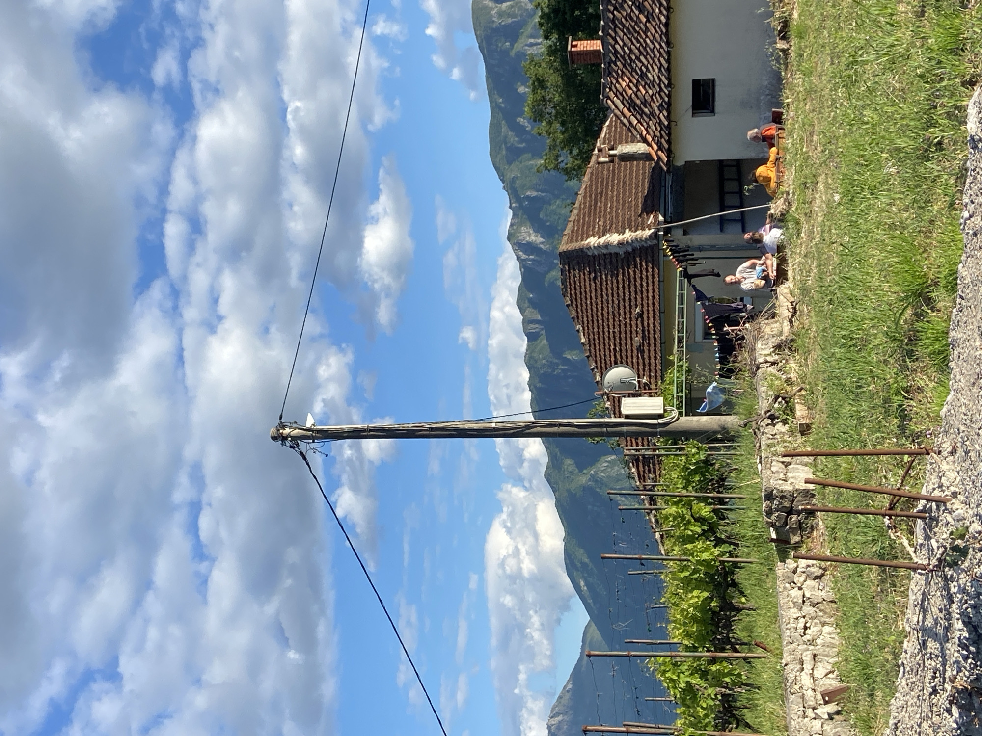 This image depicts a serene rural scene in Sotonići, Montenegro, under a bright, partly cloudy sky, likely during midday or early afternoon.
In the foreground, a tall wooden utility pole stands prominently on the left, adorned with various wires and a small white satellite dish. Power lines extend from the pole across the frame. To its right, a vineyard is visible with rows of green grapevines supported by metal stakes, bordering a rustic stone wall.
The mid-ground features a traditional, light-colored house with a distinct red-tiled roof. An outdoor patio or porch area connected to the house hosts several people. One individual wearing an orange top is partially seen behind a small outdoor table. Nearby, two other people are seated, one in a white top holding a bottle, and another person also dressed in a light-colored top. Laundry is hanging to dry near the house, indicating a domestic setting.
In the background, majestic green mountains rise, their contours visible against the sky, suggesting a mountainous or hilly region. The sky above is a vibrant blue, dotted with numerous fluffy white cumulus clouds.
The scene conveys a sense of peaceful rural life. No visible text is present in the image.