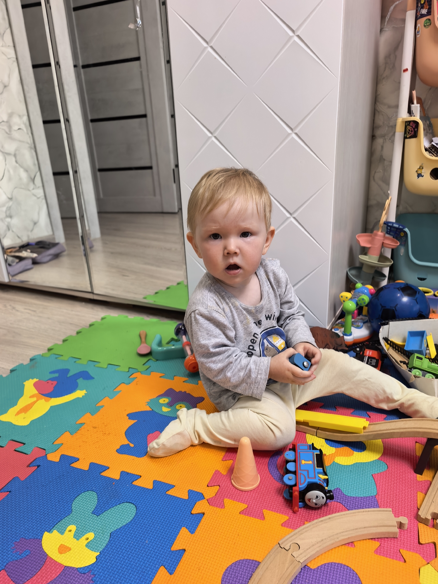The image depicts a young Caucasian child, appearing to be around one to two years old, sitting on a colorful interlocking foam mat on the floor. The child has light blonde hair and is wearing a grey long-sleeved shirt and light-colored pants. The child is holding a small blue toy block in their right hand. The mat is a jigsaw puzzle of various colors, including green, blue, orange, and pink, and features cartoon animal figures. Scattered around the child are various toys, including a blue toy train, wooden train tracks, and other play items that are difficult to identify precisely. In the background, a white closet with a textured geometric pattern is visible. To the left of the child, a large mirrored wardrobe reflects the room, showing a doorway and part of the floor. The overall environment appears to be a child's playroom or bedroom. The specific location within Sterlitamak, Russia, cannot be determined from the visual information provided.