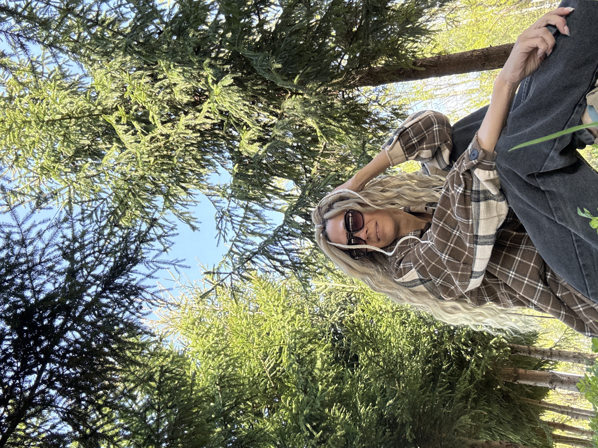 A low-angle outdoor shot depicts a woman situated among tall green trees beneath a blue sky. The woman is wearing a brown and white plaid flannel shirt and dark-colored jeans. She has long blonde hair and sunglasses. The trees surround her, and the sky is visible between their branches. The location is Unknown.