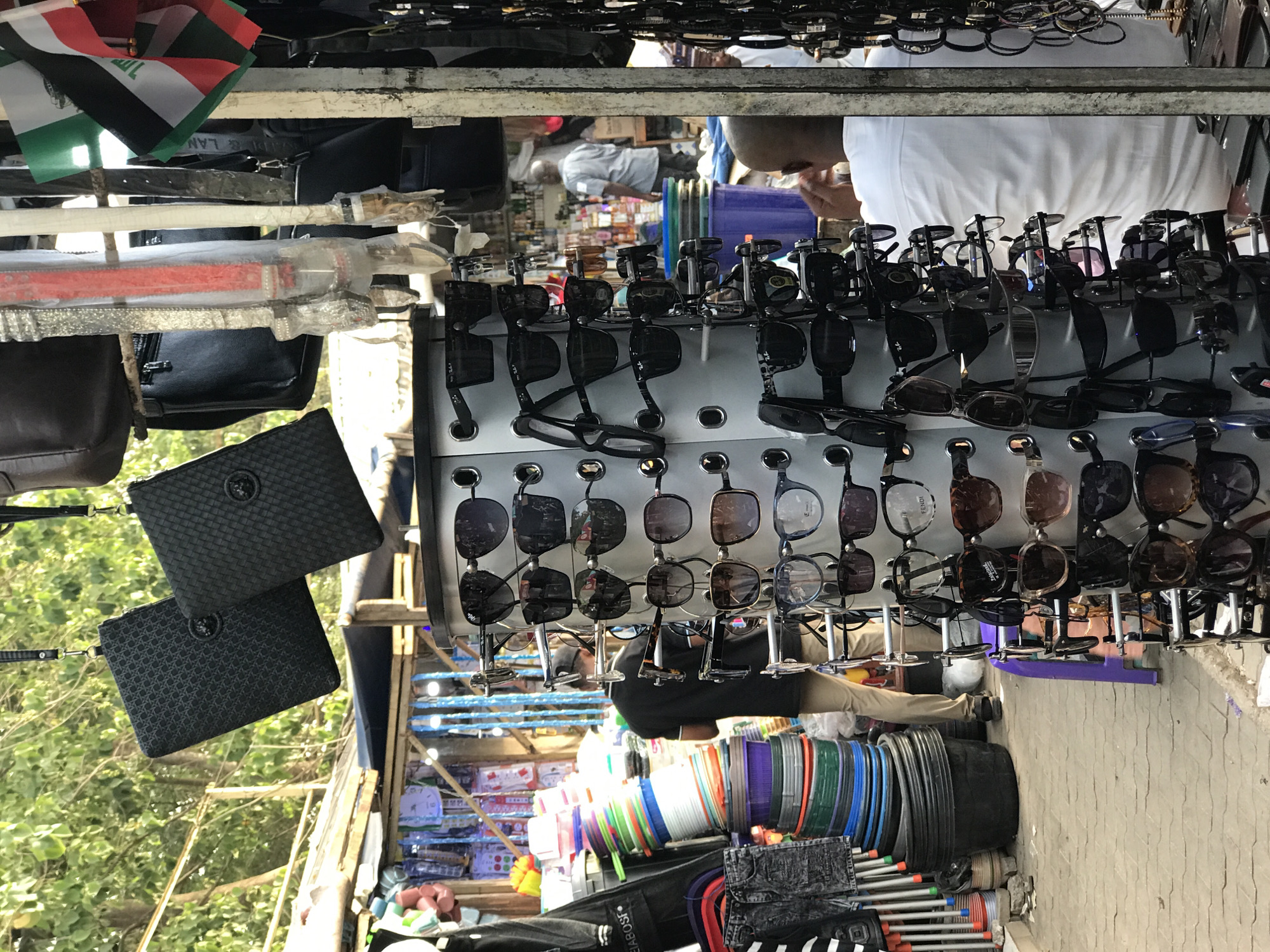 This image captures a vibrant market stall scene, likely outdoors or semi-outdoors, given the visible foliage and natural light. The setting, identified as Abuja, Nigeria, features a diverse array of goods displayed for sale.
In the foreground and center, numerous pairs of sunglasses are meticulously arranged on a multi-tiered, rotating display stand. These appear to be fashion accessories, with some visible text on the frames or lenses, including "FENDI" and "BALMAIN," suggesting branded items. Above the sunglasses, a collection of black handbags or purses are hanging, two of which are prominent: one with a woven texture and another with a repeating patterned design, both featuring a circular metallic emblem. Further up, what appear to be decorative or ceremonial swords or long knives, wrapped in clear plastic, are suspended. A small flag, possibly the Iraqi flag with red, white, black, and green stripes and Arabic script, is also visible at the top left.
Several individuals are present, though mostly partially obscured. One person wearing a white shirt is visible from the chest up, seemingly interacting with the stall or customers. Another individual in a dark top and tan trousers is visible from the waist down, appearing to be crouched or seated behind the display of sunglasses. A reflection of a person in a white shirt can also be seen in the lenses of some sunglasses, adding to the sense of activity.
In the lower part of the image, rows of brightly colored plastic bowls, buckets, and other containers are neatly stacked, indicating a variety of household goods on offer. The ground beneath the stall is paved with light-colored bricks or tiles. The overall impression is that of a bustling marketplace during daylight hours, with vendors presenting their merchandise. Other items like packaged goods on shelves are discernible in the background, further reinforcing the market environment. Visible text also includes "SEABOUSE" on a black item at the bottom.