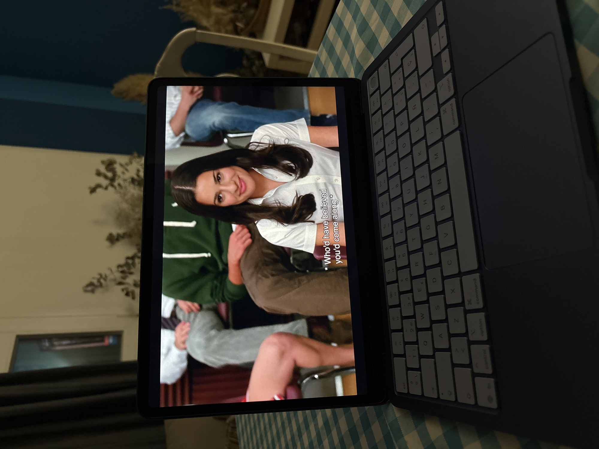 The image captures a tablet, likely an iPad, displaying a video, positioned on a blue and white checkered tablecloth. The tablet is attached to a dark-colored keyboard case, which is open to the right.
On the tablet screen, a woman with dark hair, wearing a white collared shirt, is prominently featured, smiling towards the viewer. She appears to be an actress in a scene from a TV show or movie. Parts of other people are visible around her on the screen, including one individual in a green top and another in grey. Subtitles are displayed on the lower right of the screen, reading: "* Who'd have believed you'd come along *".
The keyboard case shows a light grey QWERTY key layout, including Spanish characters (e.g., 'Ñ'). Visible keys include 'control', 'option', 'command', 'shift', 'tab', 'esc', 'caps lock', 'fn', and various letter, number, and symbol keys. A dark trackpad is integrated into the right side of the keyboard.
The background is dimly lit, suggesting an indoor setting during the evening or night. Blurred elements include furniture, such as a chair, and indistinct decorative items that resemble plants. The scene takes place in Urbanización Encinares De Alcolea, Spain.