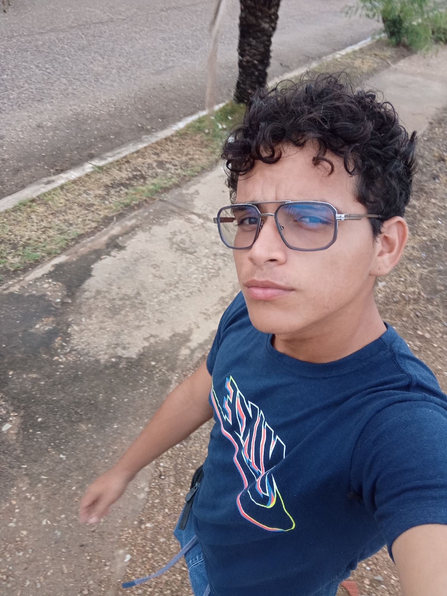 A medium shot taken outdoors shows a young male of Hispanic descent from a slightly elevated angle. He has curly, dark hair and is wearing blue-tinted eyeglasses with silver rims. His eyes are open, and he is looking towards the camera with a neutral expression. He is wearing a navy blue t-shirt with a graphic design in white, red, blue, and yellow that resembles a stylized wing or flame.
The background consists of a paved surface, possibly a sidewalk or path, with patches of dried grass and dirt. To the left, there is a darker, wet-looking area on the pavement. In the upper left and center of the background, a narrow strip of asphalt road is visible, with some scattered pebbles. A palm tree trunk is partially visible in the upper center-right of the frame. The lighting suggests it is daytime.
The man is standing and appears to be moving, as indicated by the slight blur in his left arm and hand, which is extended slightly forward. His right arm is bent at the elbow, and his hand is not visible. The lower portion of his body shows he is wearing denim jeans. The overall scene is one of a casual, outdoor setting in a city environment, specifically identified as Juan Griego, Venezuela.