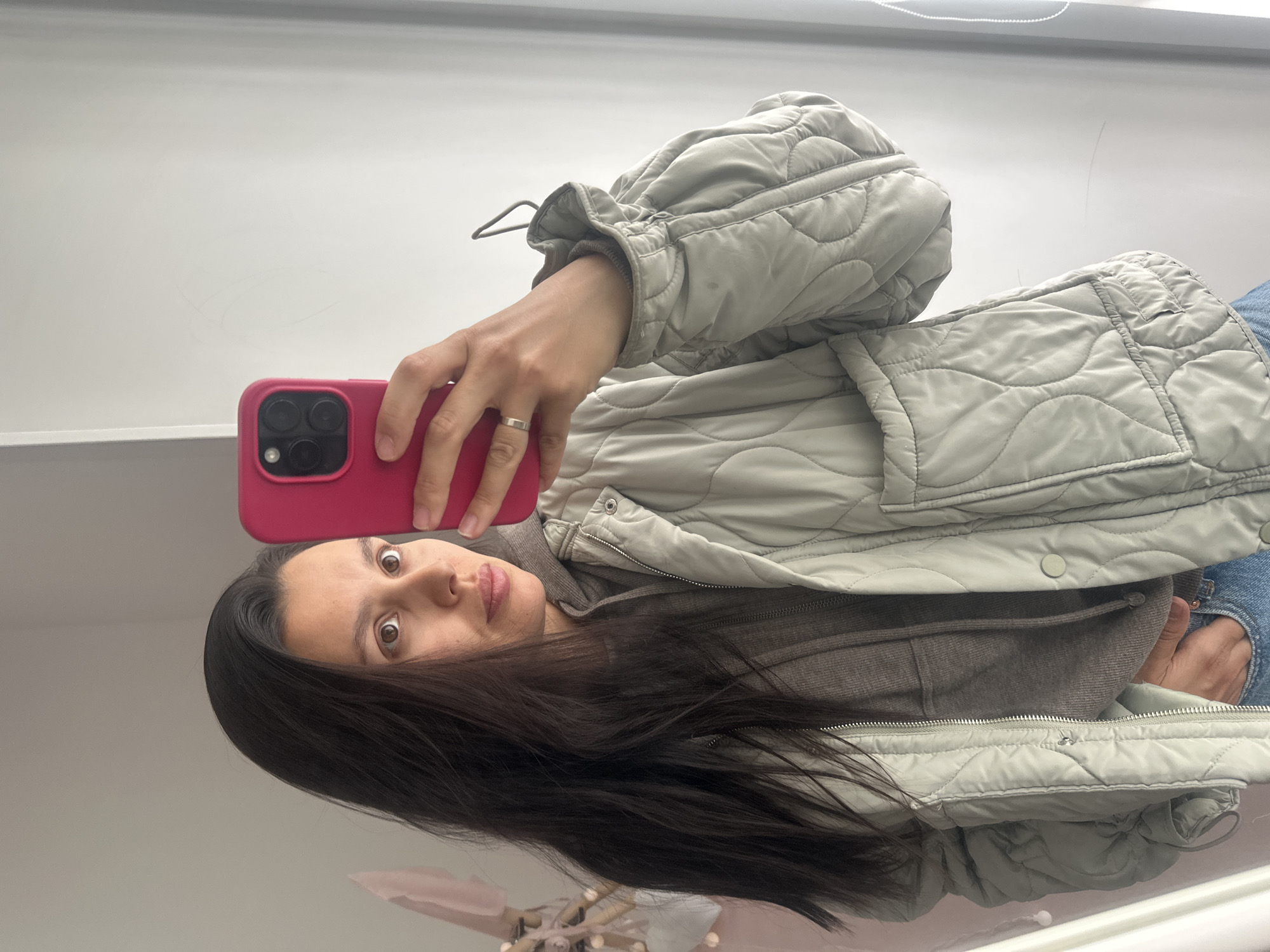 A woman with long, dark hair is taking a selfie in what appears to be an indoor location. She is holding a pink smartphone up in front of her face. She is wearing a gray, quilted jacket over a gray sweatshirt and jeans. The background is predominantly white. It is not possible to determine if this image was taken in the city of Chaikovsky, Russia.
