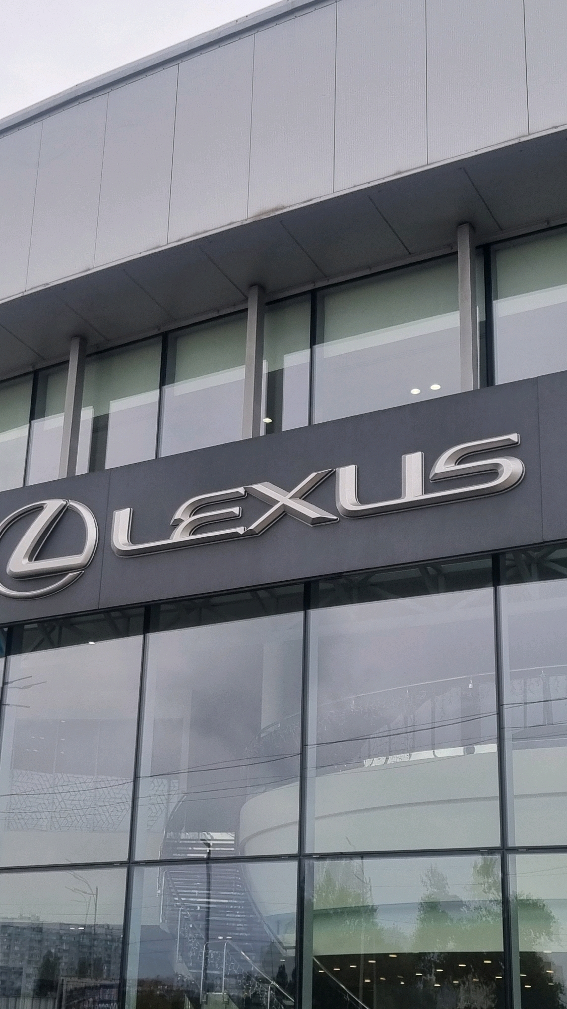 The image shows the facade of a Lexus dealership in Sofiivska Borshchahivka, Ukraine. The building features a modern architectural design with extensive use of glass and grey metallic panels. Prominently displayed on the dark grey facade is the silver Lexus logo, which consists of an oval shape with a stylized "L" inside, followed by the word "LEXUS" in raised silver lettering. The building has multiple floors with large, reflective glass windows that show reflections of the sky, trees, and interior architectural elements such as a staircase. The sky above is overcast. The scene is captured from a low angle, looking upwards at the building.