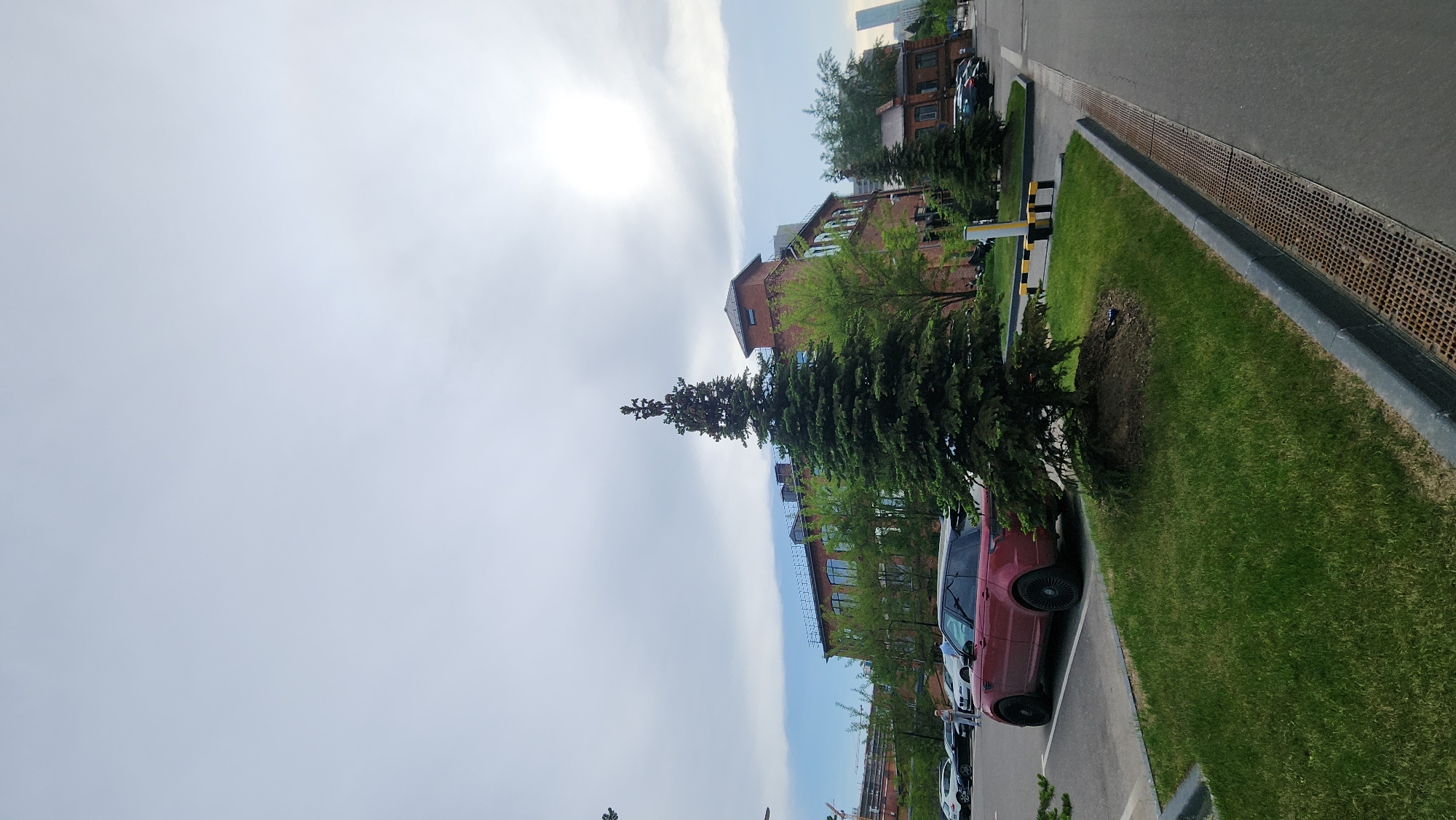 The image shows a view along a street in Moscow, Russia. The sky is bright with the sun obscured by clouds. Buildings are visible on one side of the street, primarily constructed of brick. A row of parked cars is adjacent to a narrow strip of grass that is bordered by a street with a grated drain running along the edge. Green trees are planted along the street, adding a natural element to the urban environment. There are no people immediately visible in the frame.