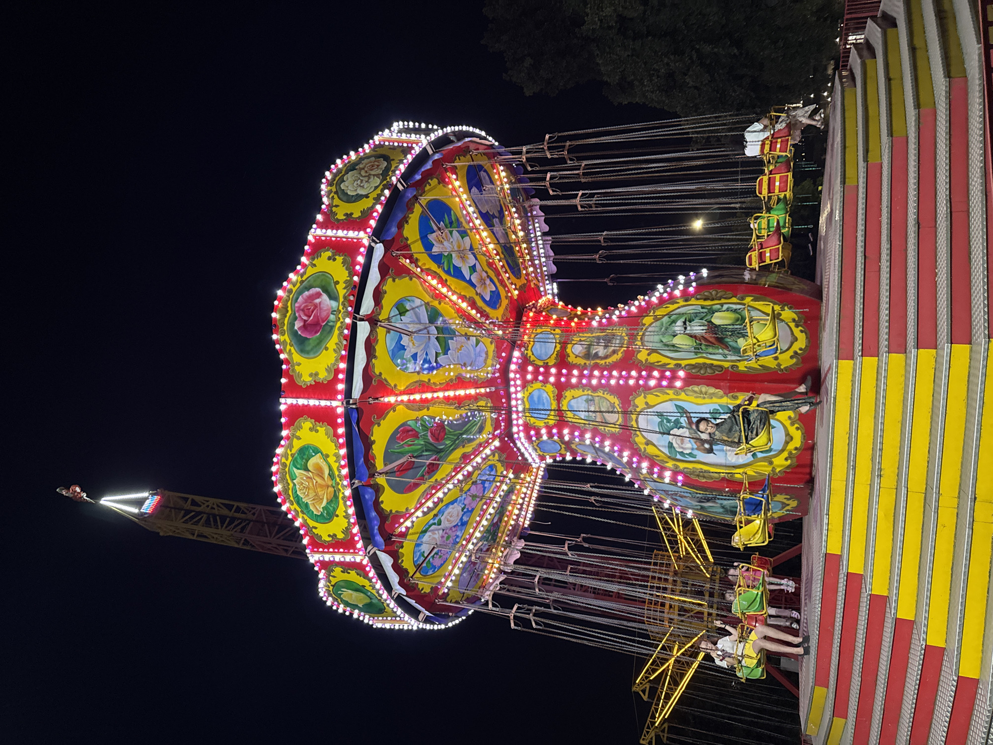 This image depicts a swing carousel ride at an amusement park in Izhevsk, Russia. The carousel is illuminated by numerous colorful lights and features ornate decorations, including paintings of flowers on its upper section. Several people are seated in the swings, some appearing to be children and others adults. The ride is situated on a raised platform with red and yellow steps leading up to it. In the background, a tall amusement ride structure, possibly a drop tower, is visible against the dark night sky. Trees are also present in the background, suggesting an outdoor setting.