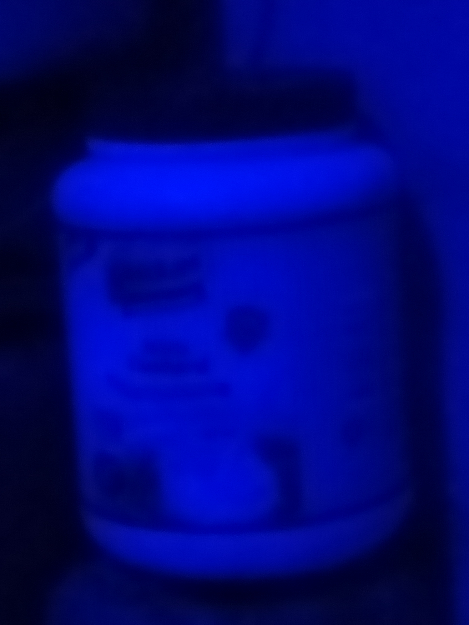 The image shows a blurred, blue-lit close-up of a white plastic container with a black lid, likely a tub of supplements or food product. The label on the container is illegible due to the blur and lighting. The container is positioned on a dark surface, possibly a shelf or table. The background is also dark and out of focus, with a dominant blue hue, suggesting artificial lighting. There is no discernible information about the location in Jalingo, Nigeria, or any human presence in the image.