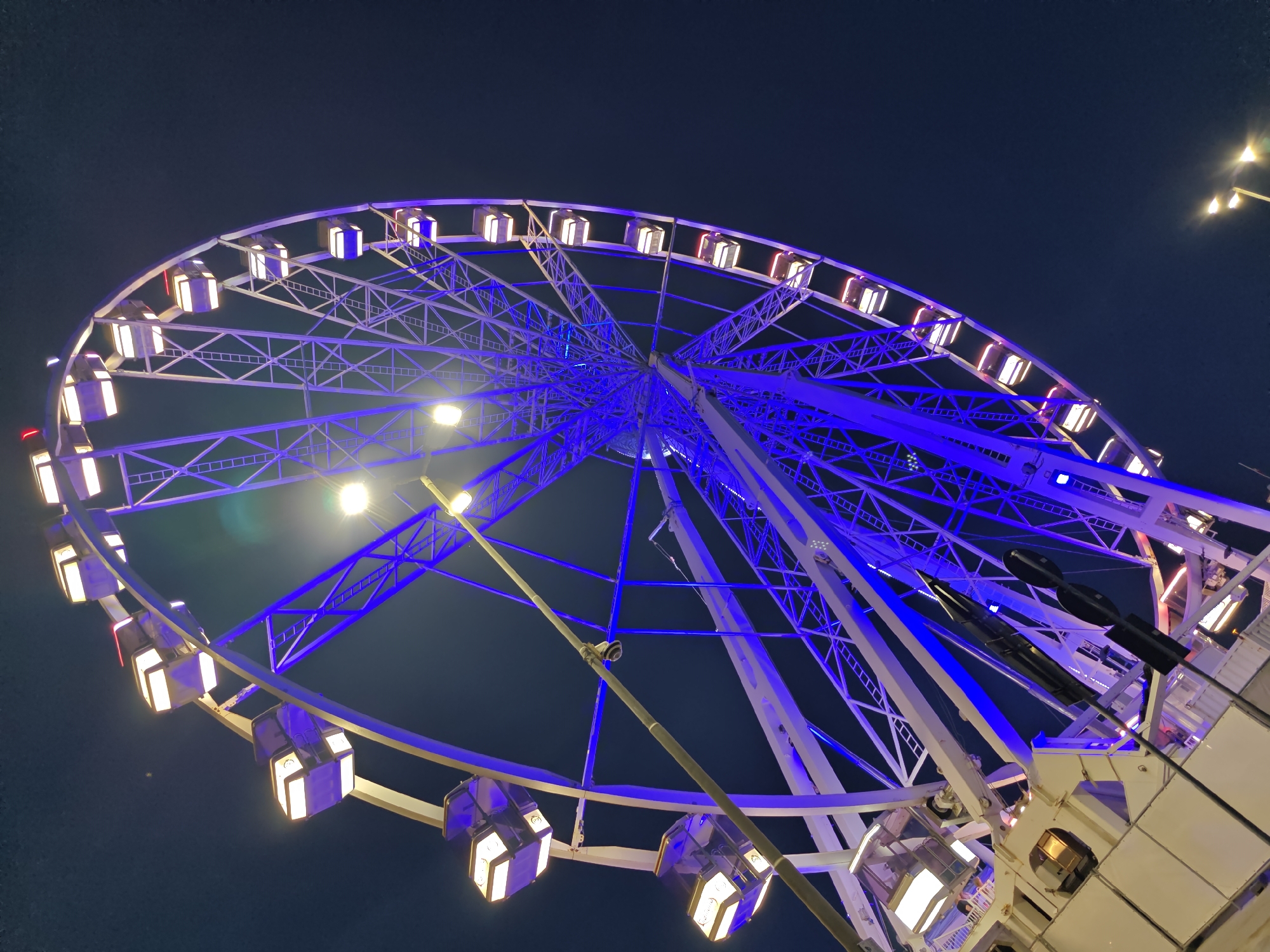 A close-up, low-angle shot captures a Ferris wheel at night in Rimini, Italy. The structure is illuminated with vibrant blue lights along its spokes and supports, while the gondolas are lit with a warm, yellow light. The dark night sky serves as the backdrop, with a few streetlights visible in the upper right corner. The Ferris wheel appears to be in operation or ready for operation, with multiple gondolas visible, some empty and others possibly occupied. The image focuses on the intricate metal framework of the wheel and the geometric patterns created by the lights. The overall impression is one of a festive and illuminated urban attraction against the darkness of the night.