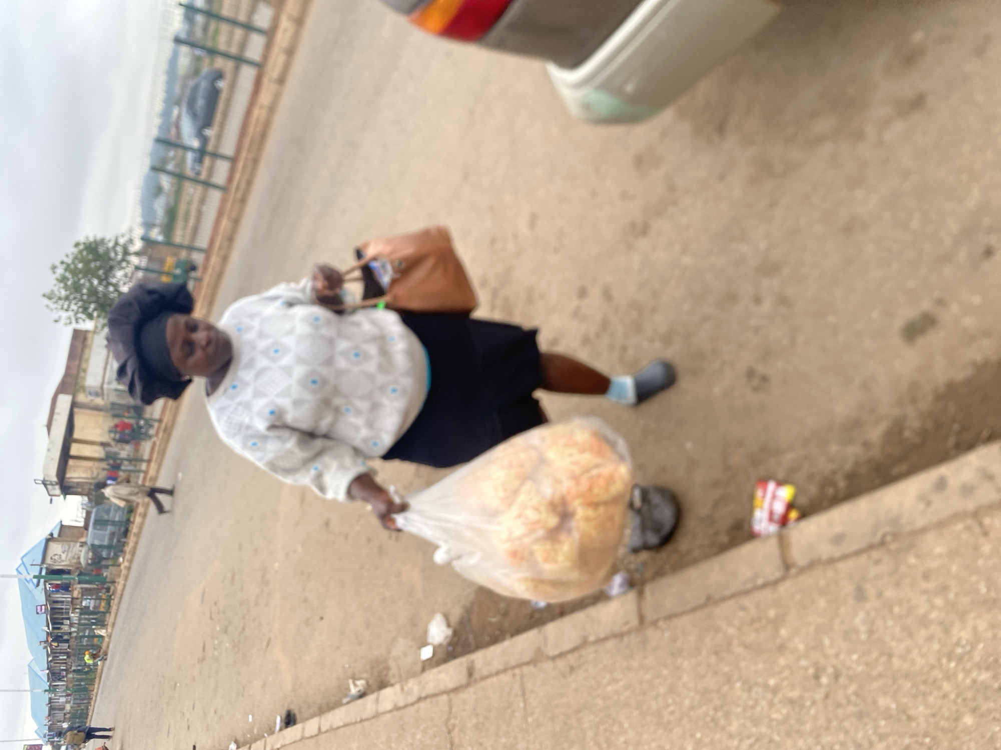A medium, slightly blurred, eye-level shot captures a woman walking along the edge of a road in Bukuru, Nigeria. The woman, dressed in a black skirt and a light-colored, long-sleeved patterned shirt, carries a large plastic bag filled with bread-like items. A brown purse hangs on her left shoulder. She wears a black head covering and is wearing grey socks with grey shoes.
The road appears cracked and worn, with litter scattered along the curb. A parked car is partially visible in the upper right corner. The background shows a busy street scene with various buildings and pedestrians. The sky is overcast.
